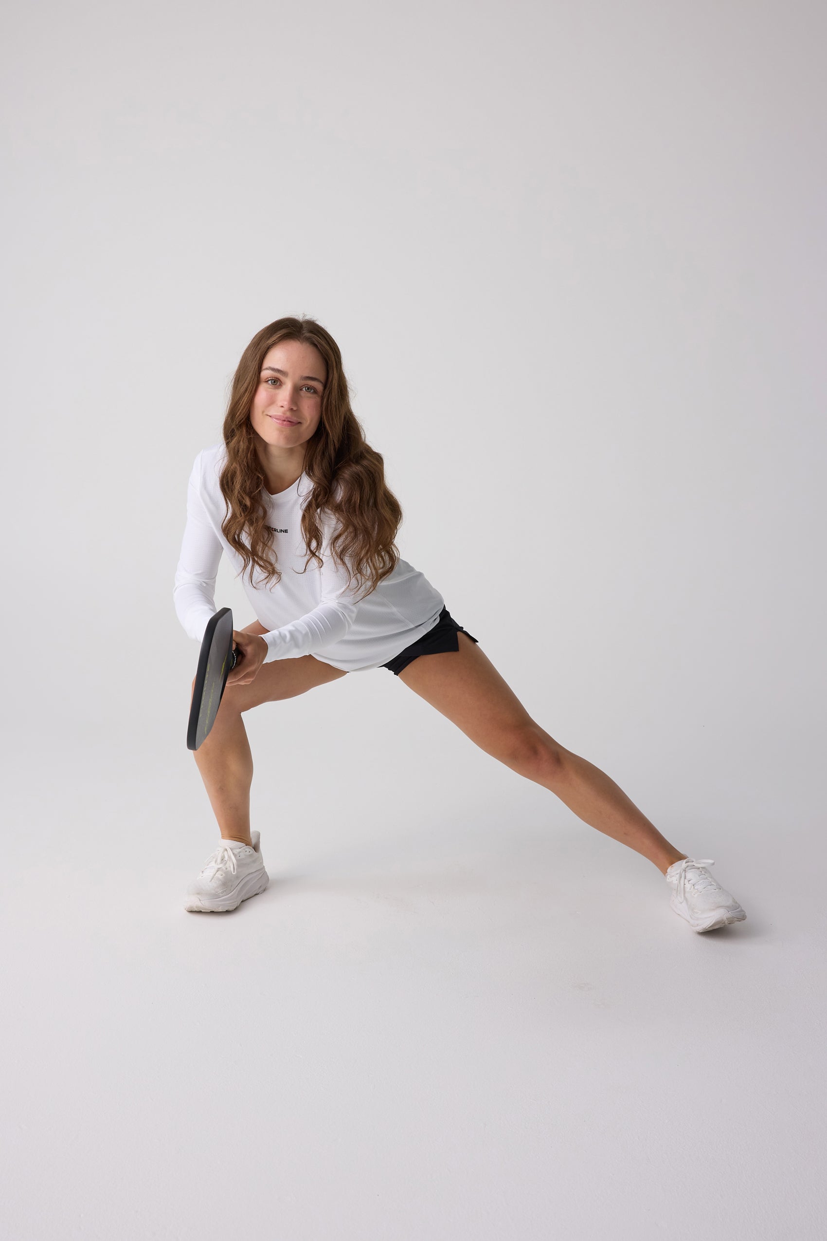 White women's long sleeve shirt by Centerline Athletics, with subtle texture and black chest logo, worn by a woman stretching in black shorts.
