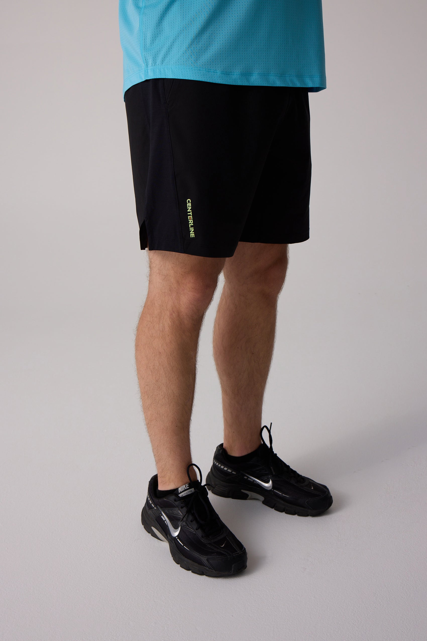 Black men's shorts by Centerline Athletics with a yellow-green "CENTERLINE" logo and side slit, worn with a blue mesh top and black athletic shoes.