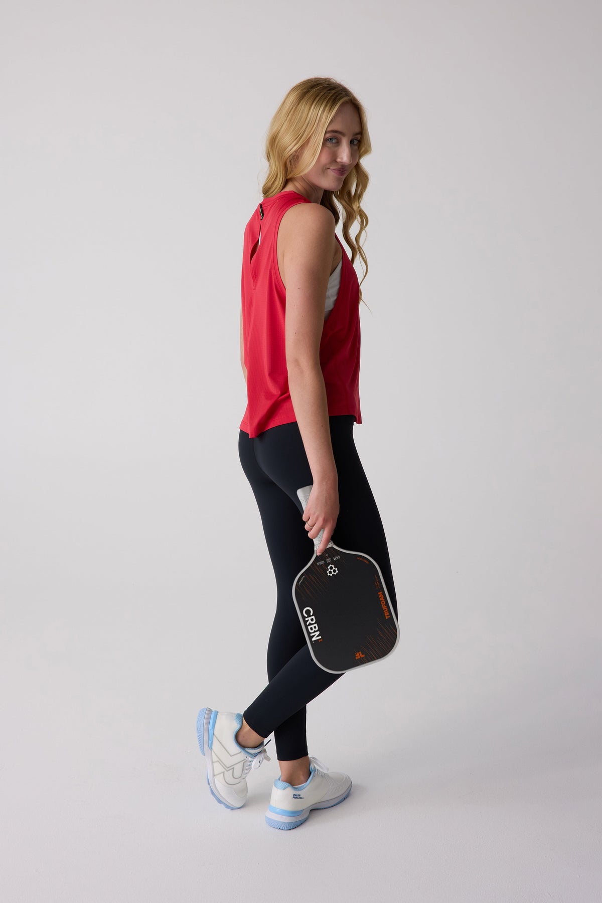 Red women's AirLite tank top by CRBN, side view with back cutout detail, worn with black leggings and white sneakers, holding paddle.
