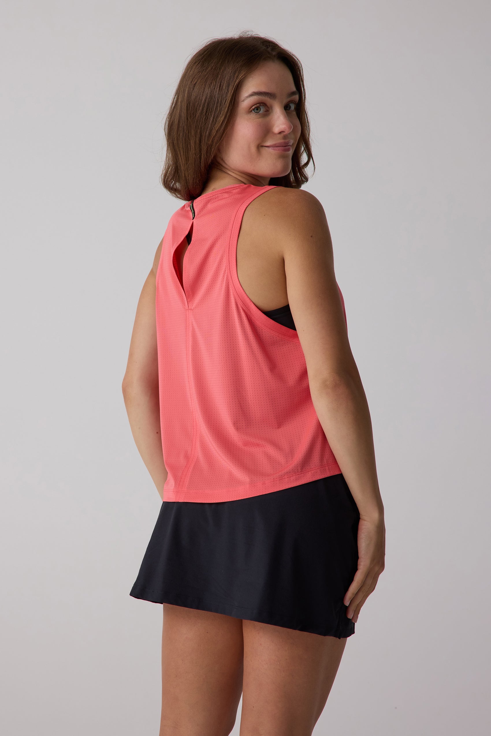 Women's coral AirLite tank top by Centerline Athletics, back view, with perforated texture and keyhole cutout, worn over a black sports bra and skirt.