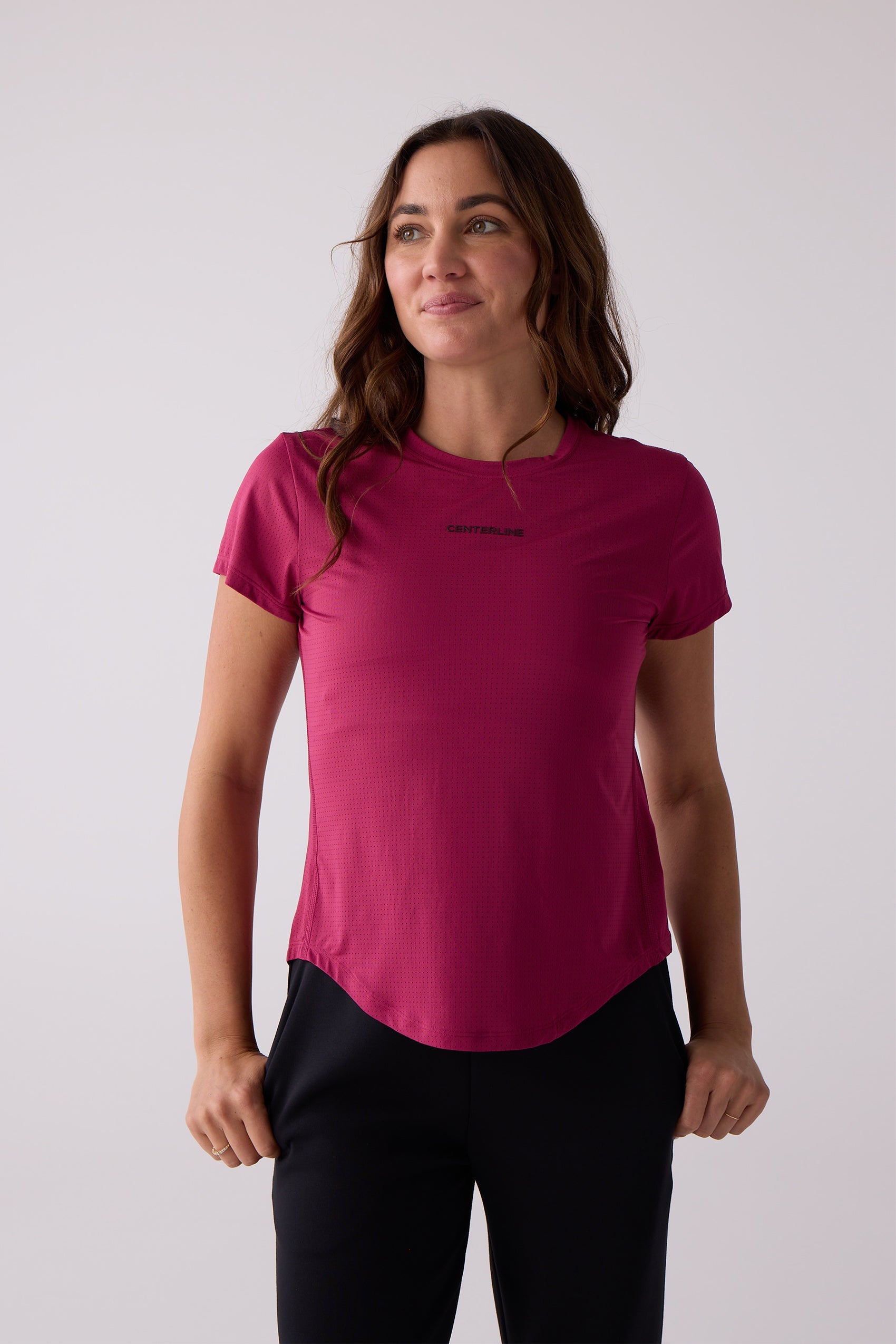 Dark pink AirLite women's short sleeve tee by Centerline Athletics with a chest logo and curved hem, worn with black pants.