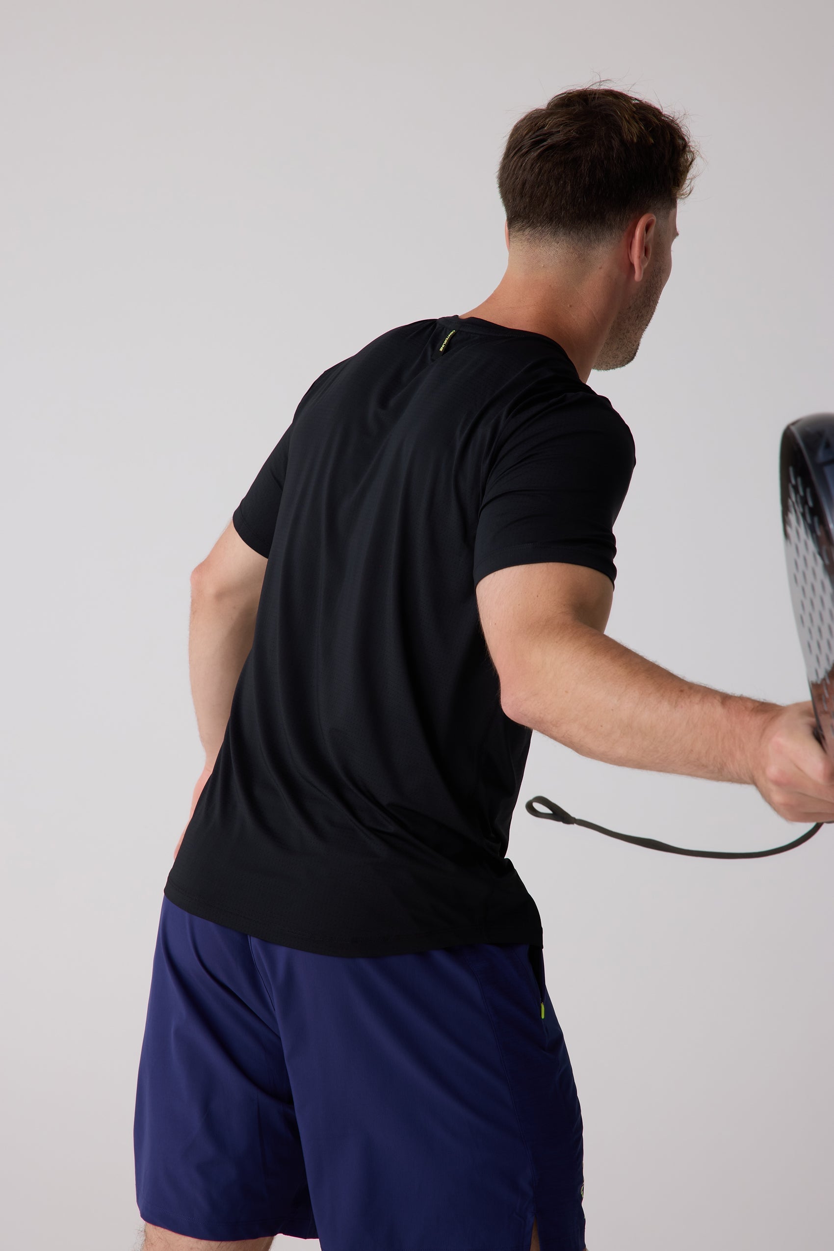 Black Men's AirLite Tee by Centerline Athletics, with subtle textured fabric, worn by a man in dark blue shorts playing paddle from the back.