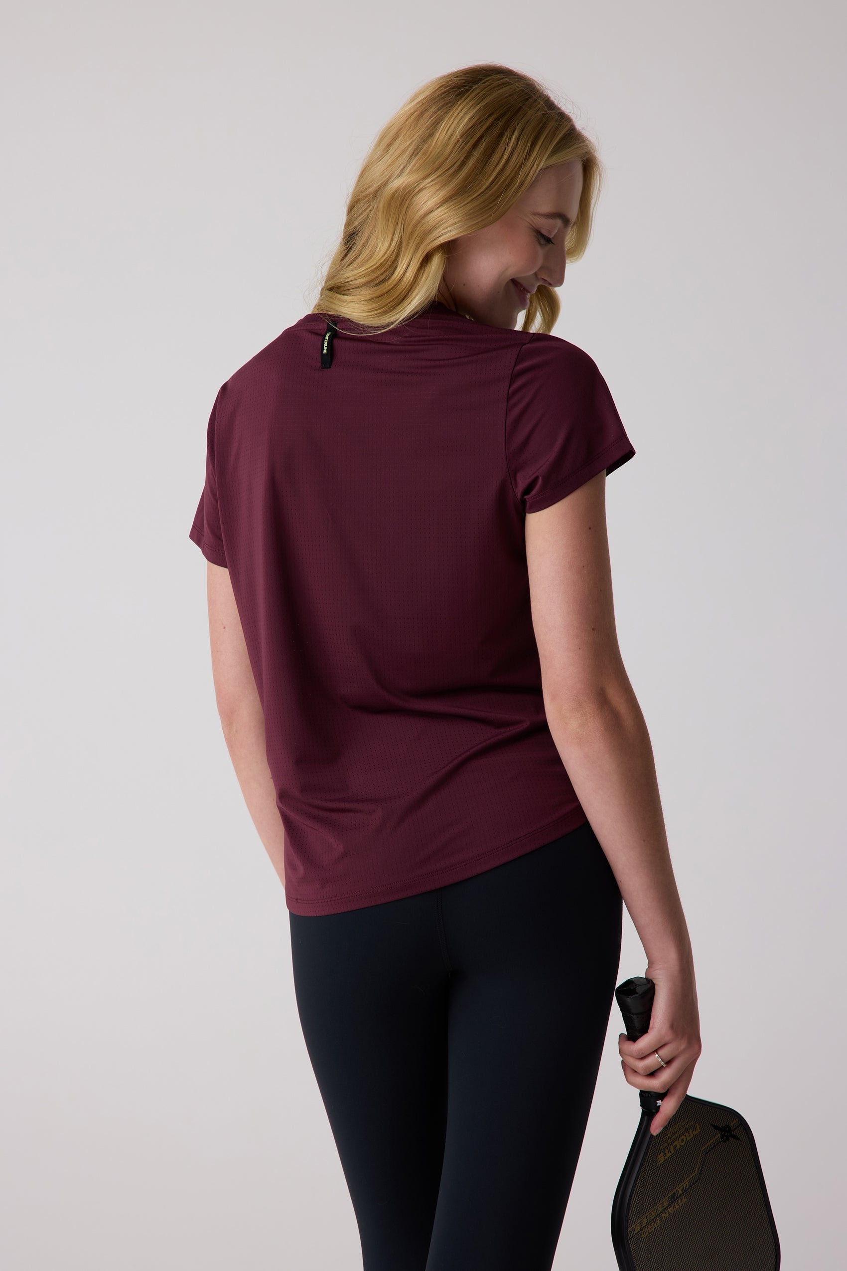 Burgundy perforated AirLite tee by Centerline Athletics with a neck tag, worn from the back with black leggings, holding a pickleball paddle.