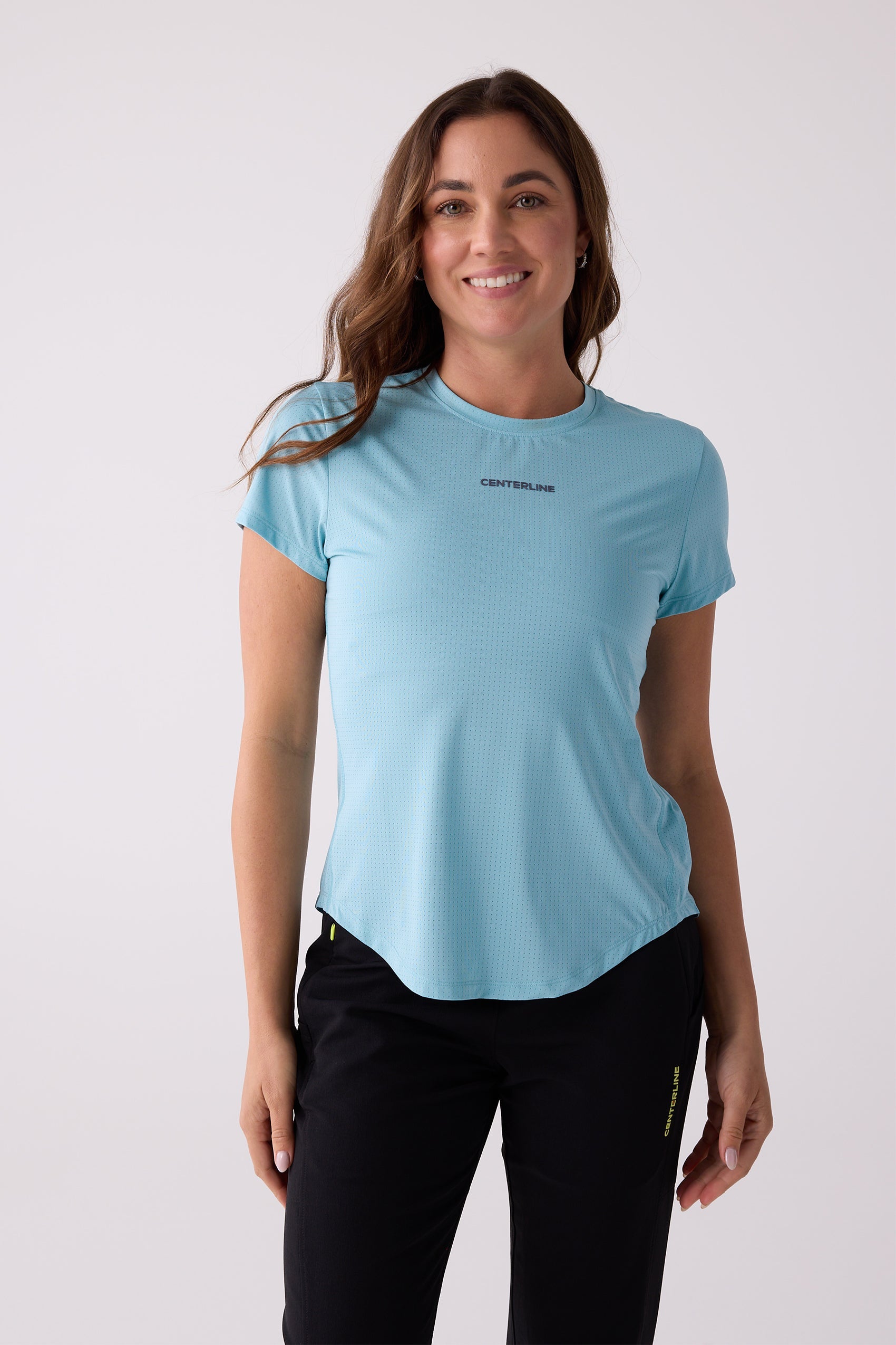 Light blue perforated women's tee by Centerline Athletics, with a crew neck and curved hem, worn by a smiling model in black pants.