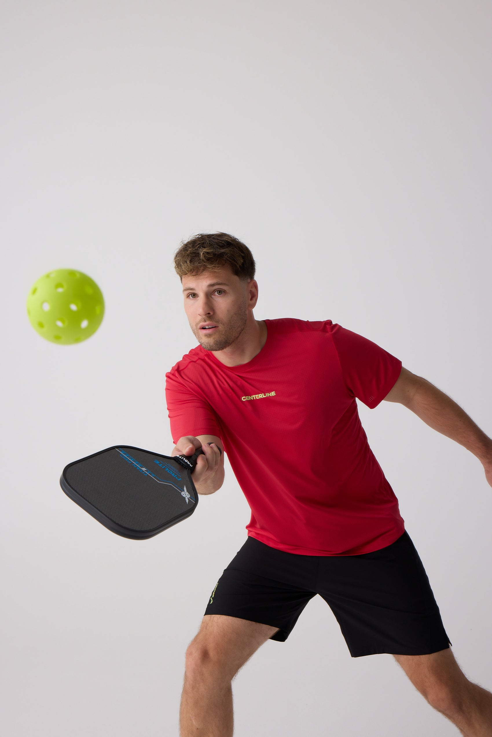 Red Men's AirLite Tee by Centerline Athletics, featuring subtle texture and a "CENTERLINE" chest logo, worn by a man playing pickleball.