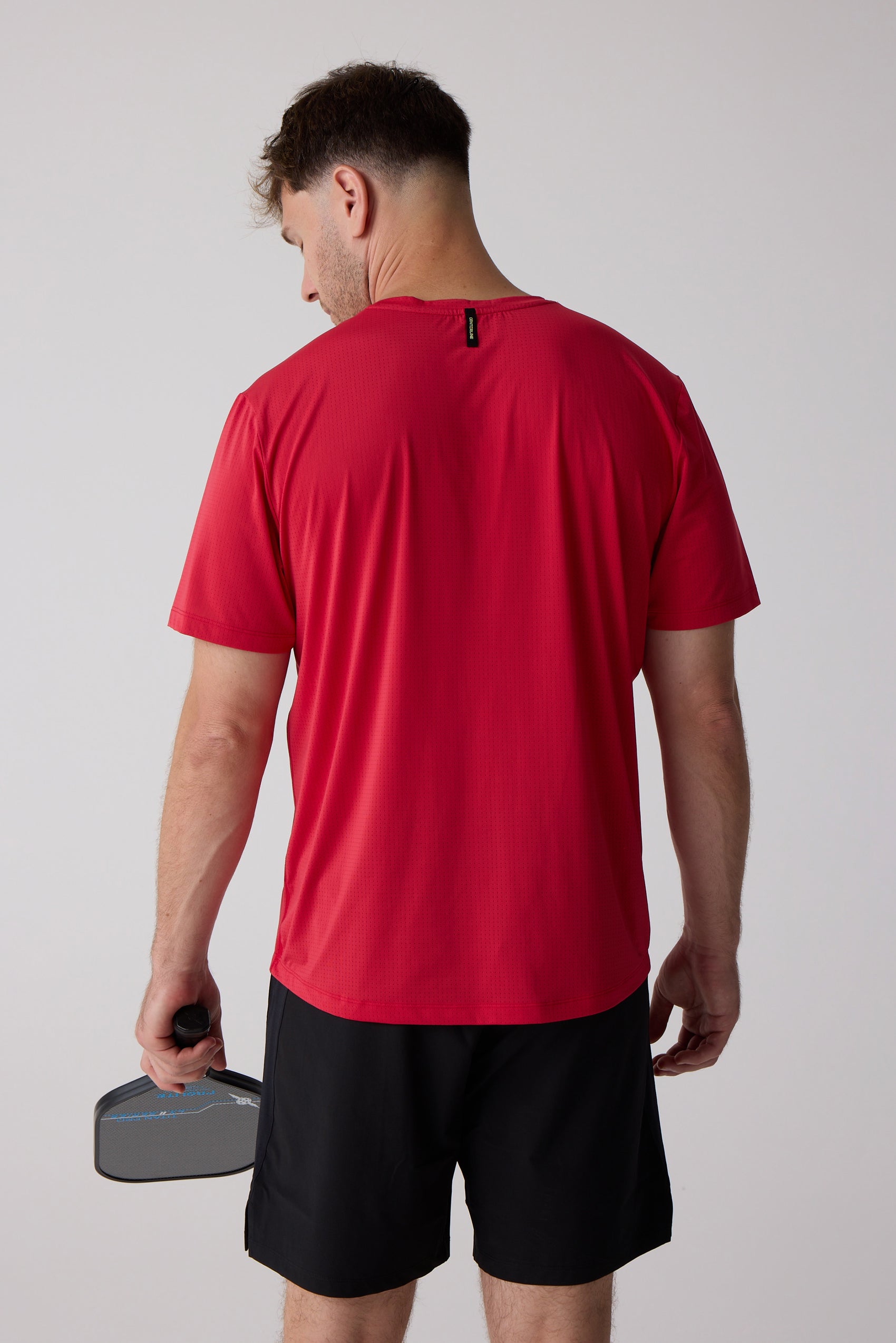 Red men's AirLite tee by Centerline Athletics with a perforated texture, shown from the back on a man holding a pickleball paddle.