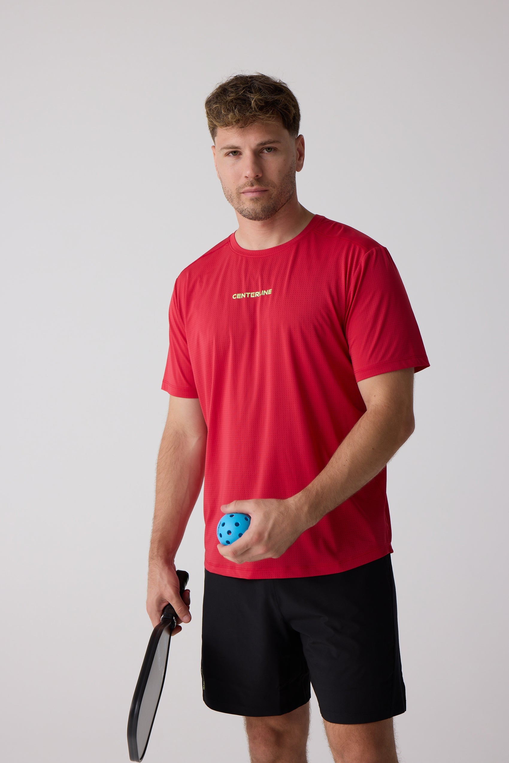 Red perforated Men's AirLite Tee by Centerline Athletics with a yellow logo, worn by a man holding a pickleball paddle and blue ball.