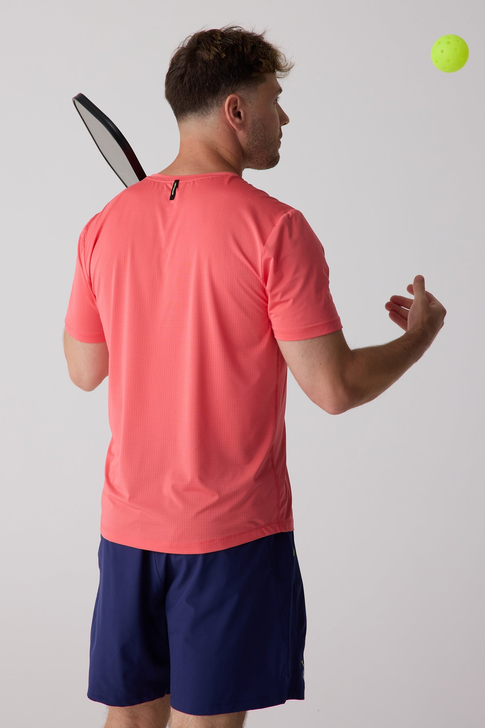 Coral short-sleeve men's AirLite tee by Centerline Athletics, with perforated fabric, worn from the back by a man holding a pickleball paddle, with a yellow pickleball in the air.