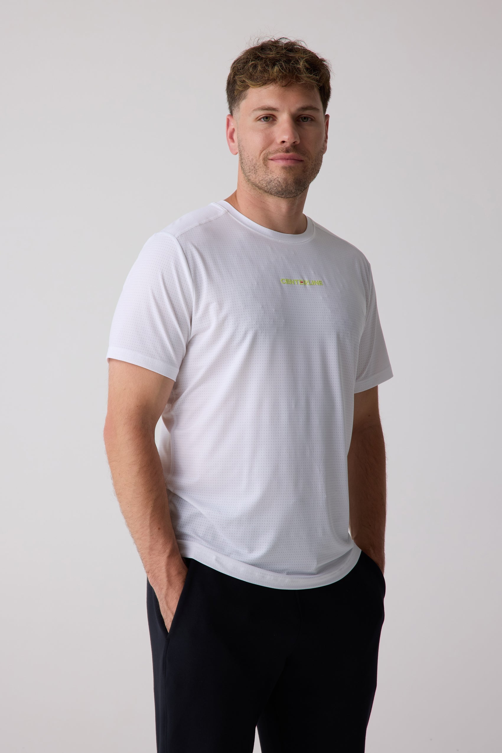 White men's AirLite tee by Centerline Athletics with perforated texture and a neon yellow logo on the chest, worn with black pants.