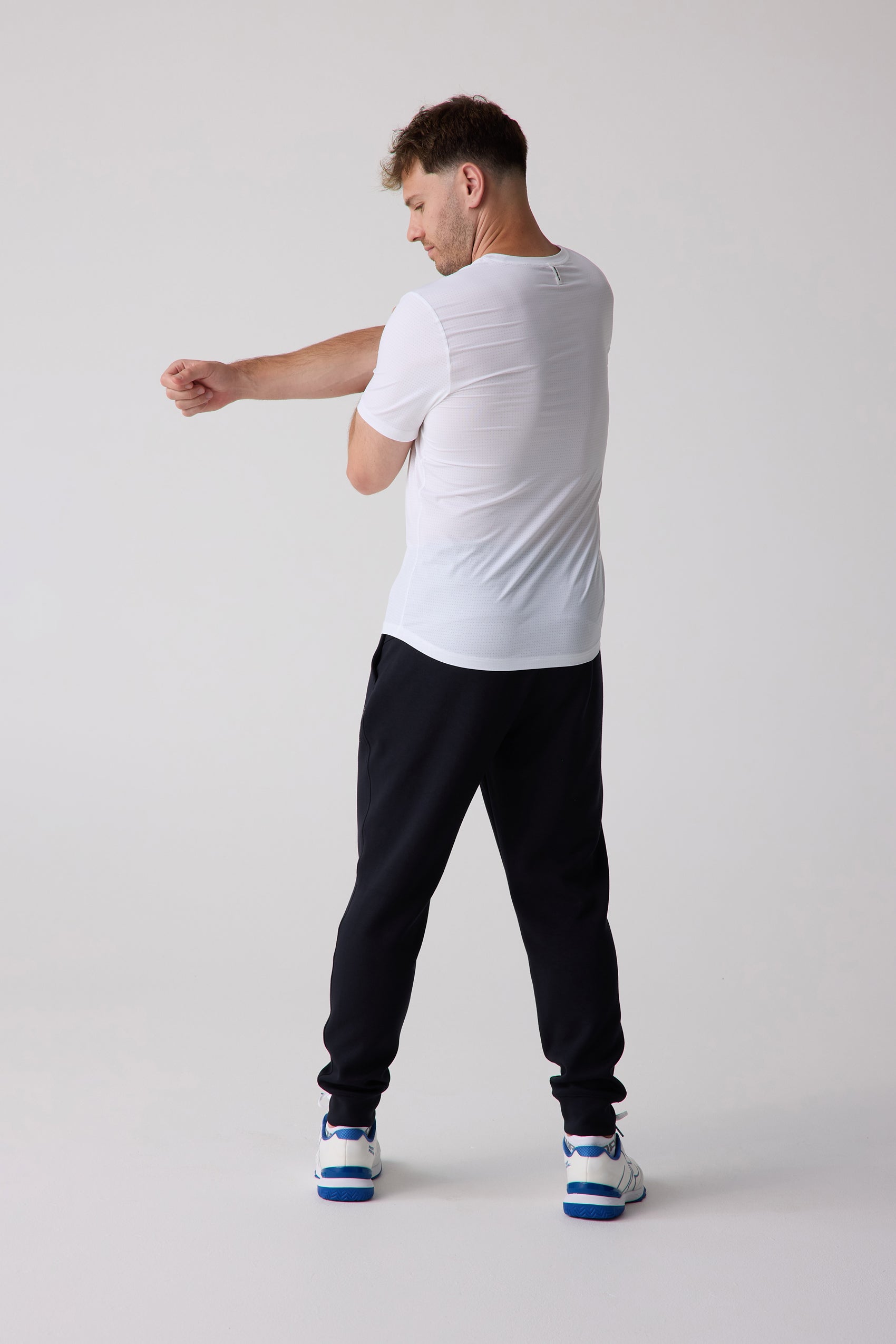 White perforated Men's AirLite Tee by Centerline Athletics, worn by a man stretching with black joggers and white-blue sneakers.
