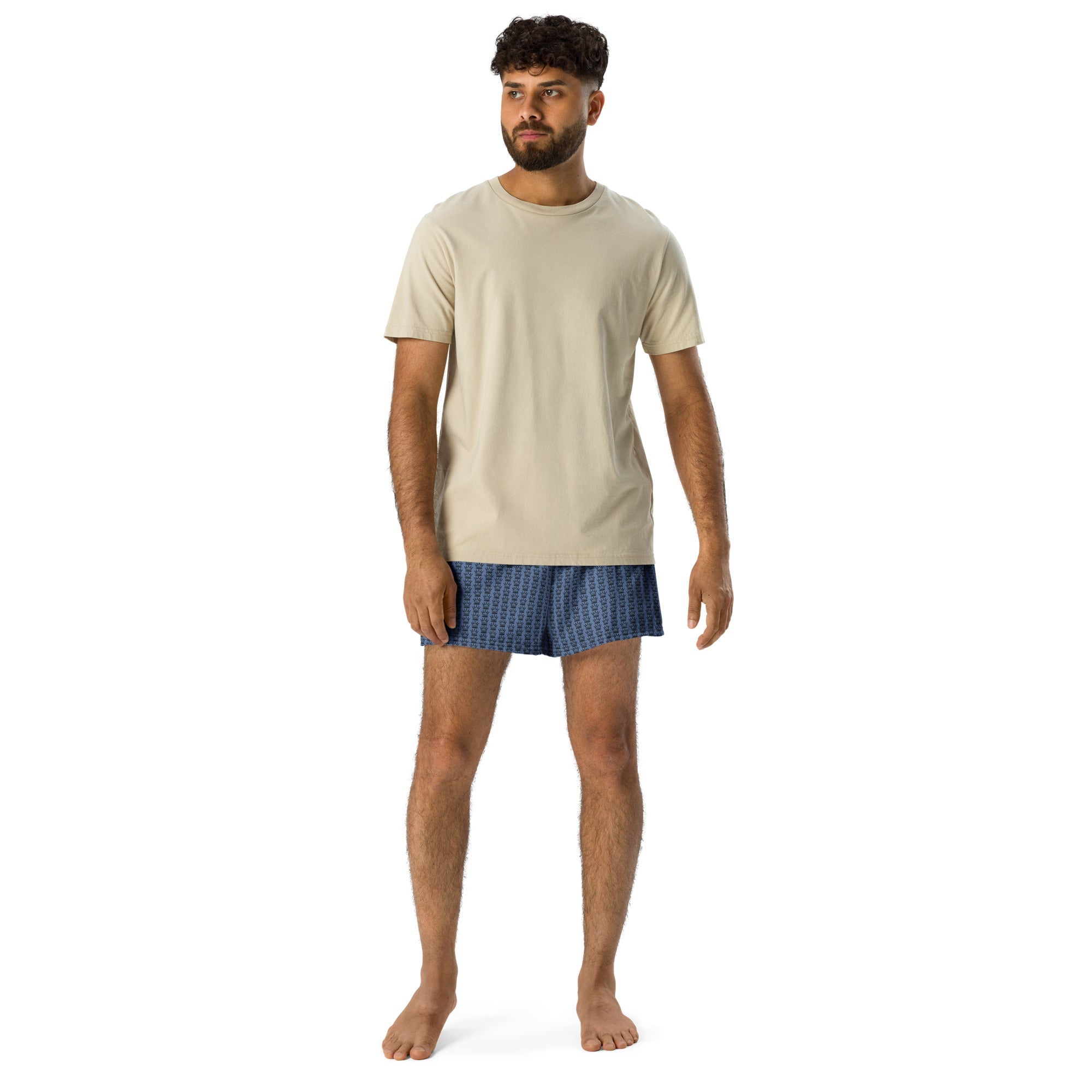Man in tan t-shirt and blue printed boxers standing barefoot on white background.
