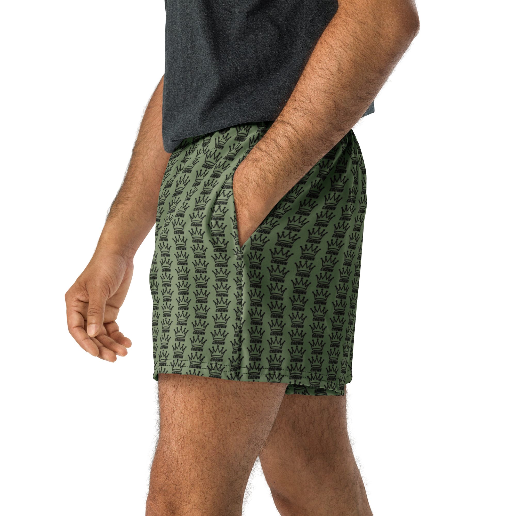Man in gray t-shirt and olive green shorts with crown pattern. One hand in pocket. Black background.