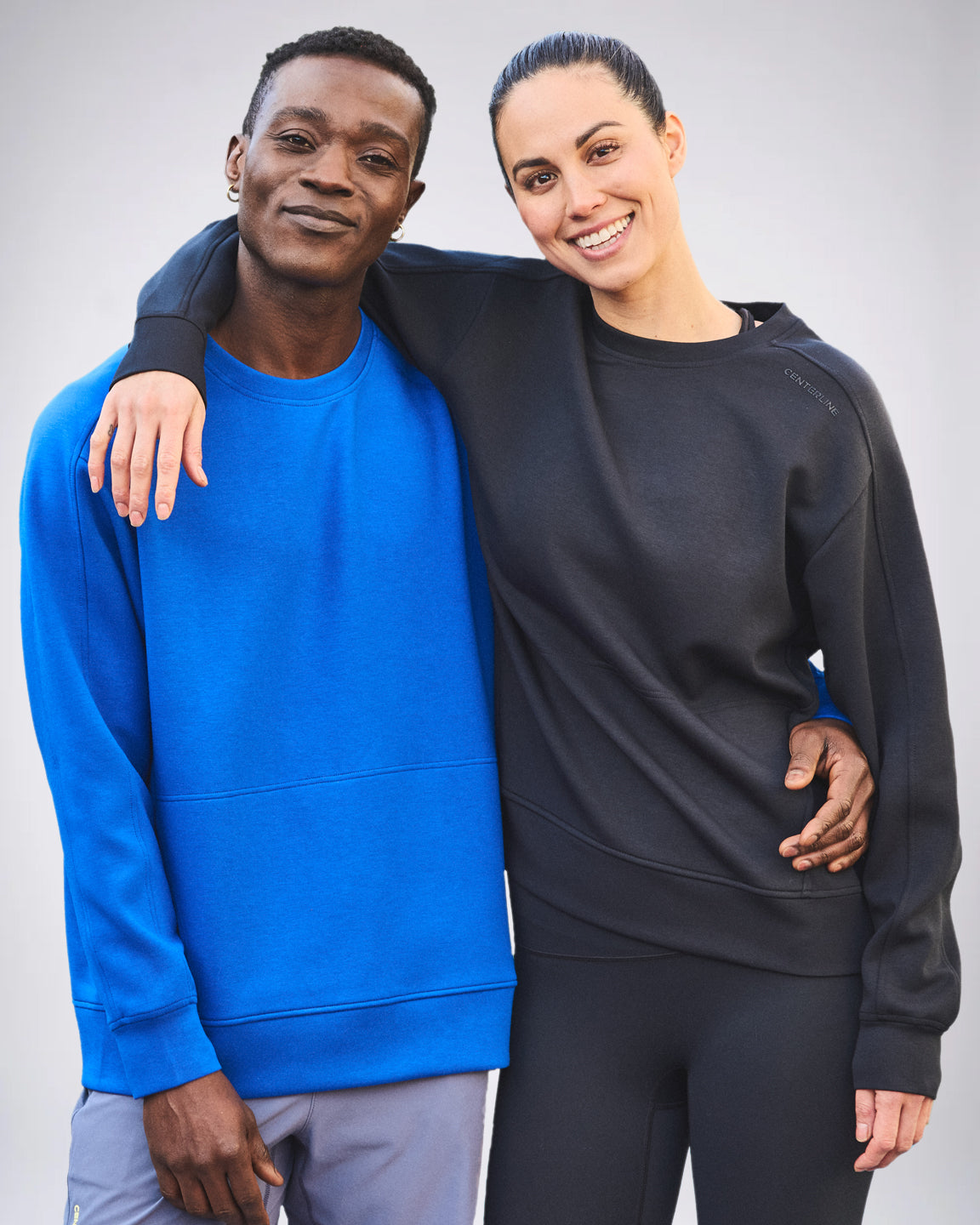 Two people in athletic wear. The man wears a blue sweatshirt and gray shorts, while the woman wears a black sweatshirt and black leggings.