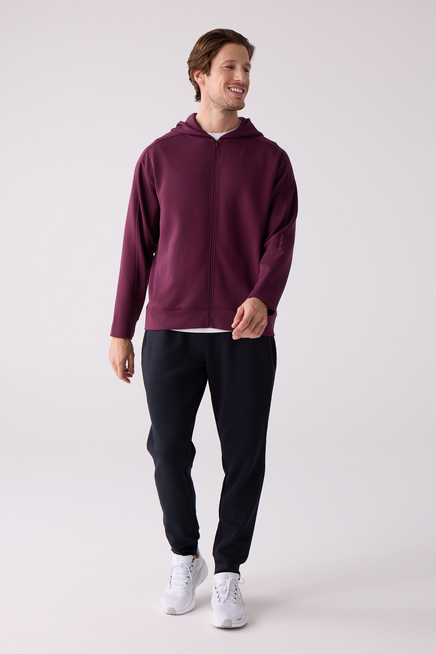 Burgundy Unisex Long Sleeve by Centerline Athletics, with a full-zip front, hood, and 'CENTERLINE' text on the sleeve, worn with dark pants and white sneakers.
