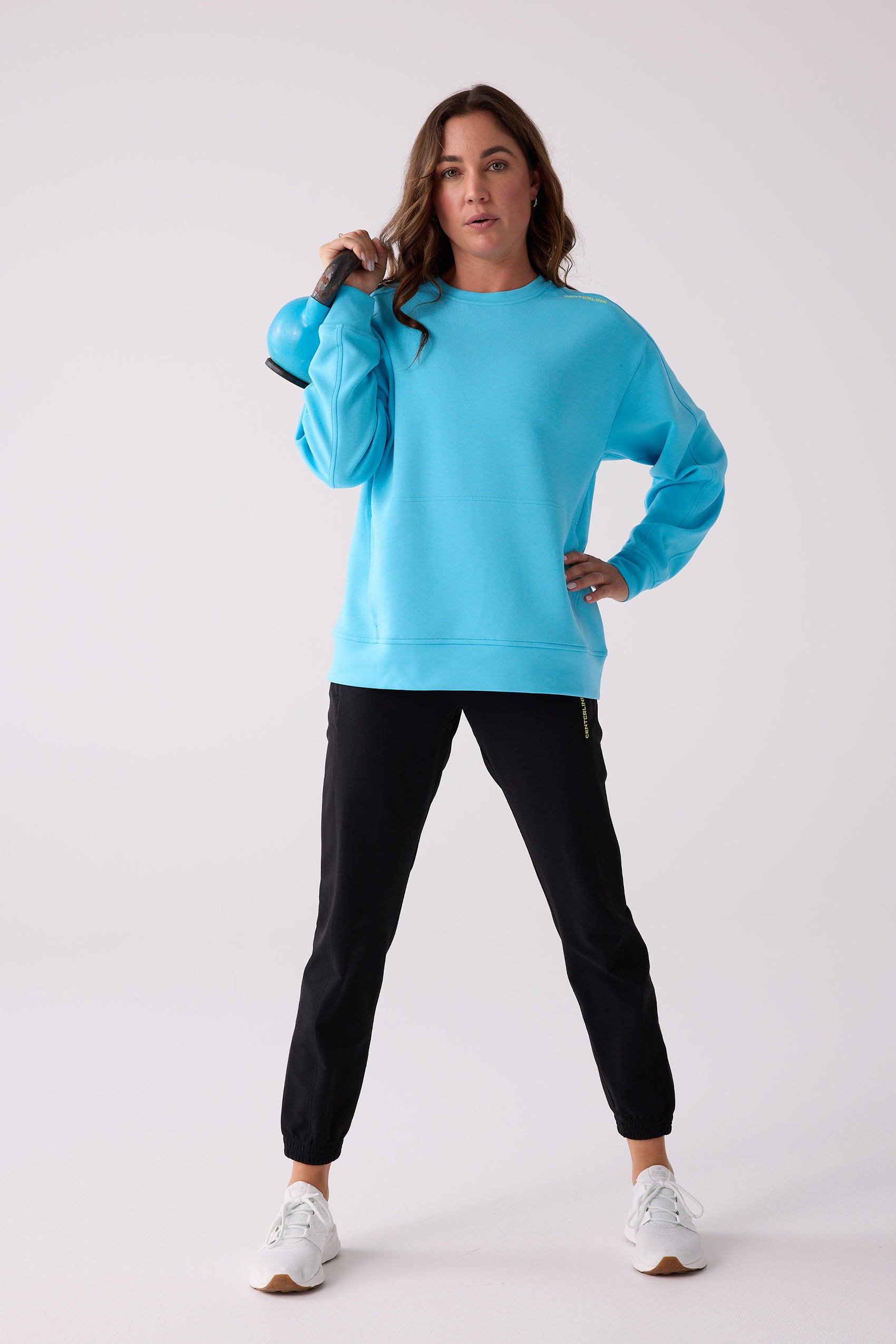 Aqua blue Women's Crew Sweatshirt by Centerline Athletics, featuring a shoulder logo, on a woman holding a blue kettlebell and wearing black joggers.