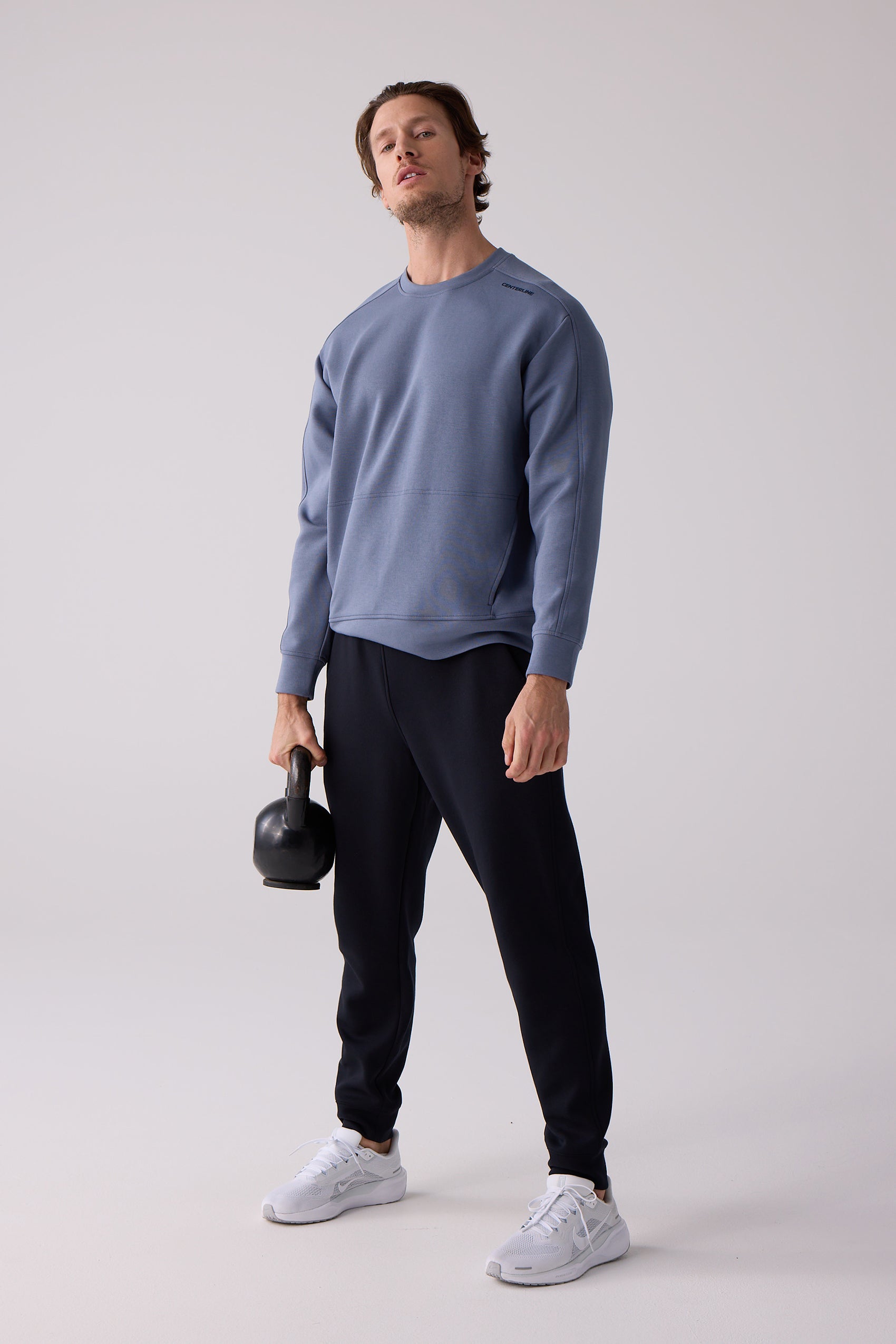 Slate blue men's crew sweatshirt by Centerline Athletics, with small shoulder text, worn by a man in black joggers and white sneakers holding a kettlebell.
