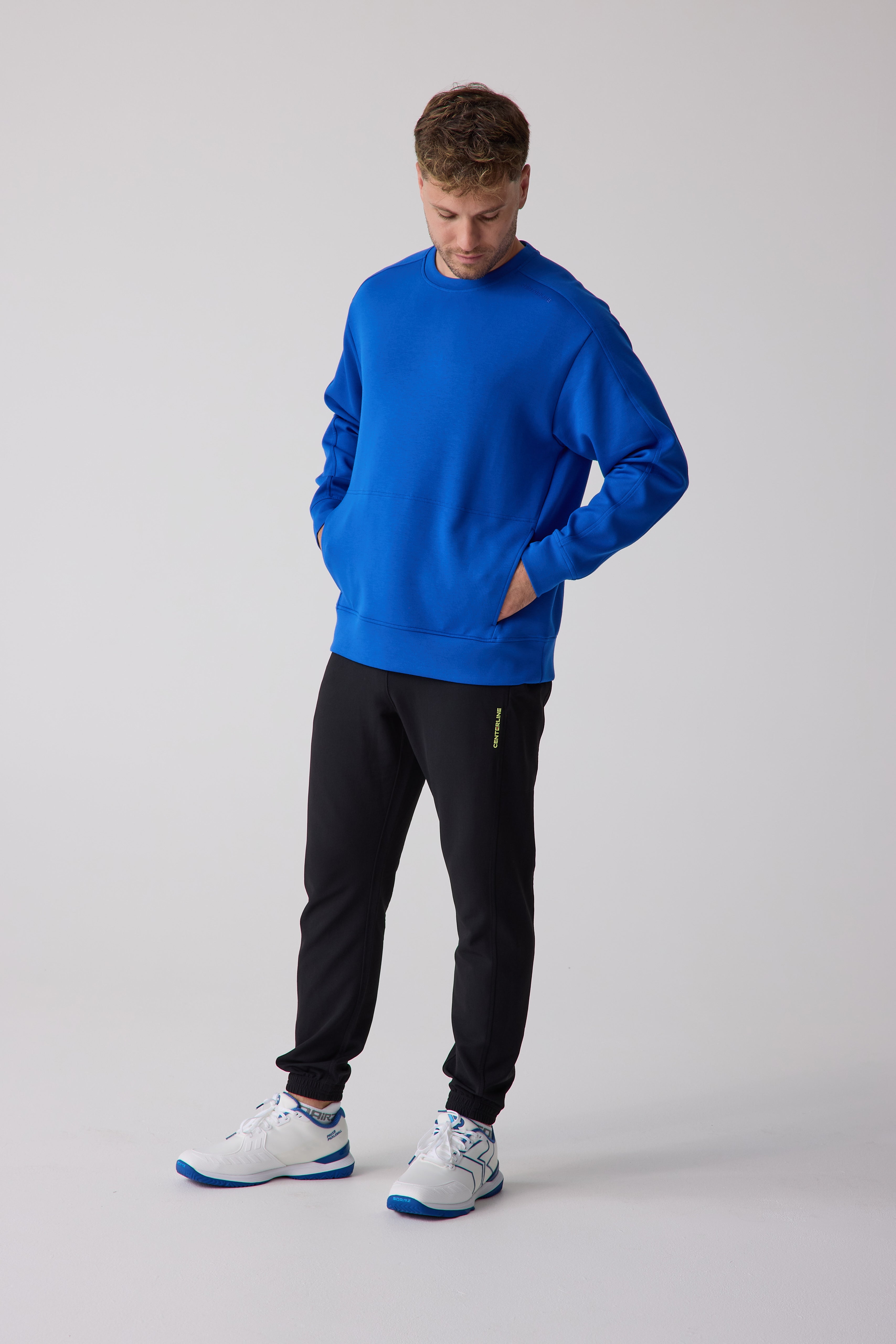Men's blue crew sweatshirt by Centerline Athletics with an embossed neck logo and front kangaroo pocket, paired with black joggers and white and blue athletic shoes.