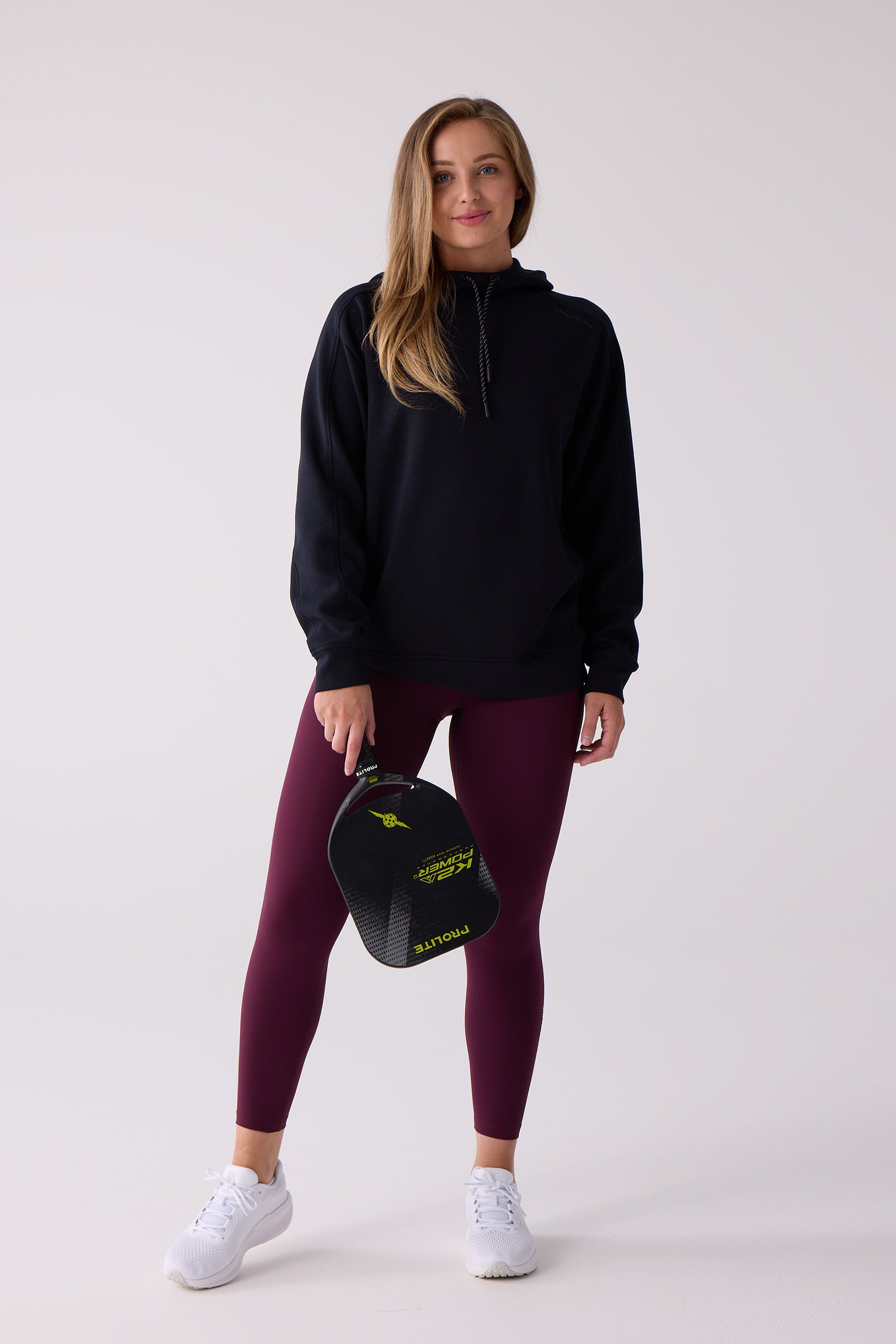 Women's black hooded sweatshirt by Centerline Athletics, styled with maroon leggings and white sneakers, holding a pickleball paddle.