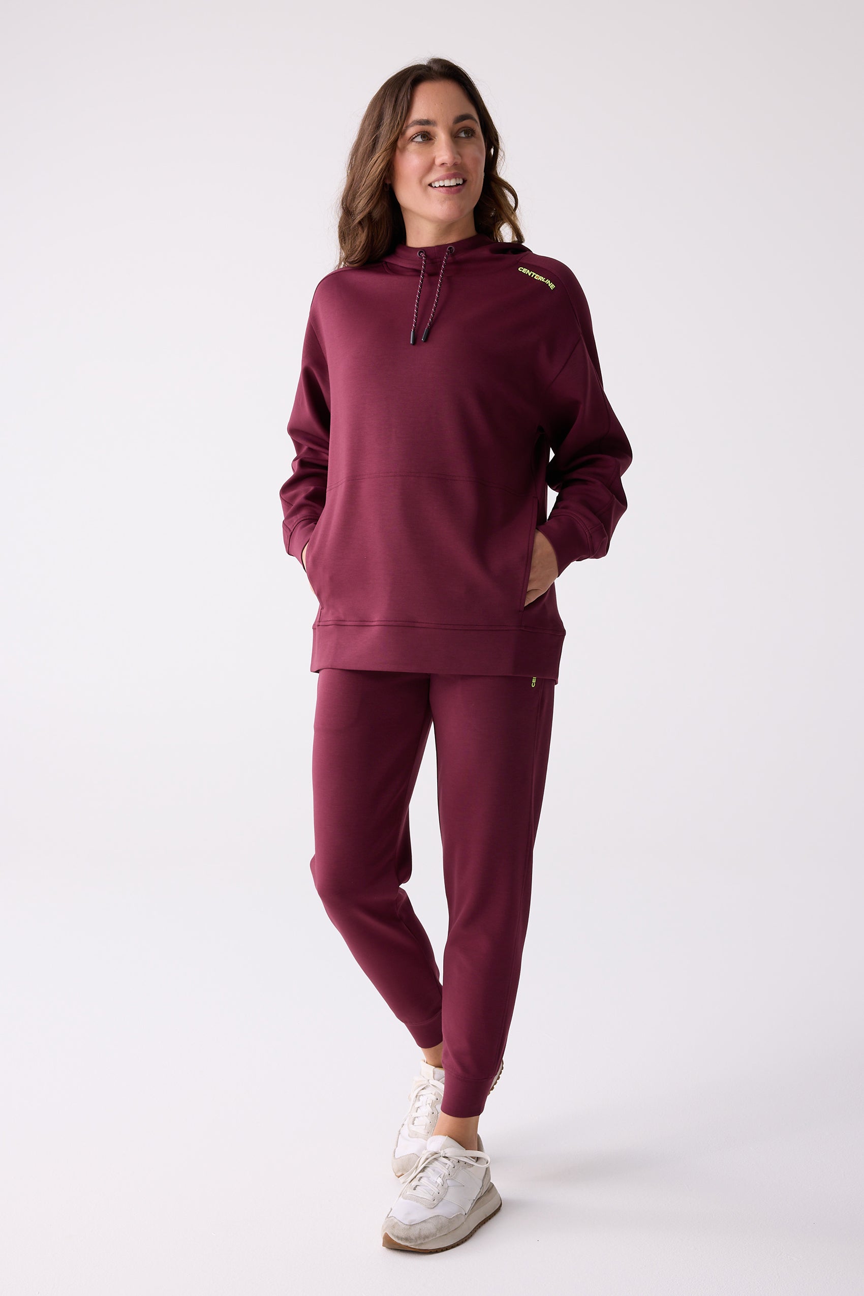 Burgundy women's hooded sweatshirt by Centerline Athletics, with neon yellow 'CENTERLINE' shoulder text, drawstring, and kangaroo pocket, styled with matching jogger pants and white sneakers.