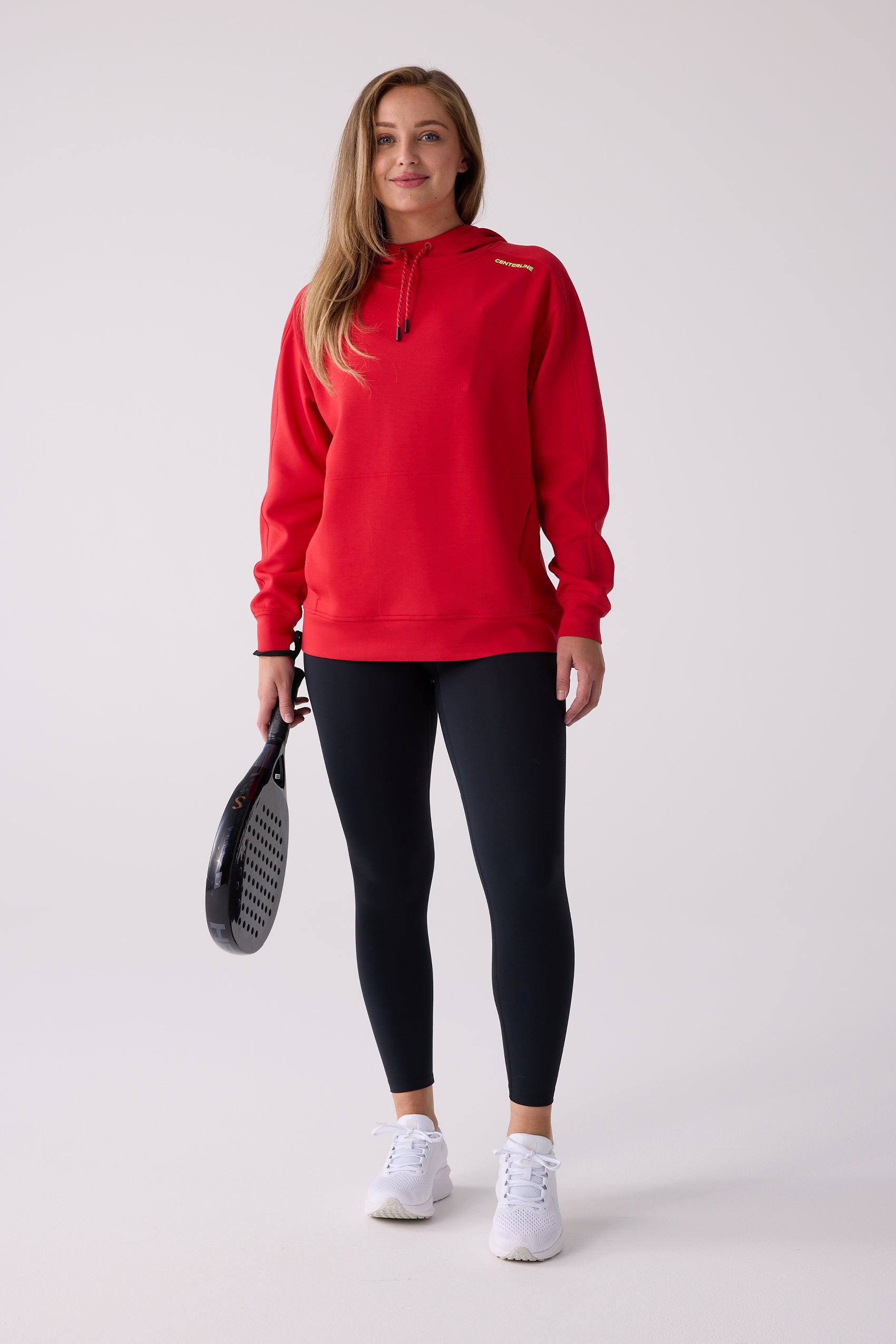 Red hooded sweatshirt by Centerline Athletics with yellow 'CENTERLINE' text, worn with black leggings, white sneakers, and holding a padel racket.