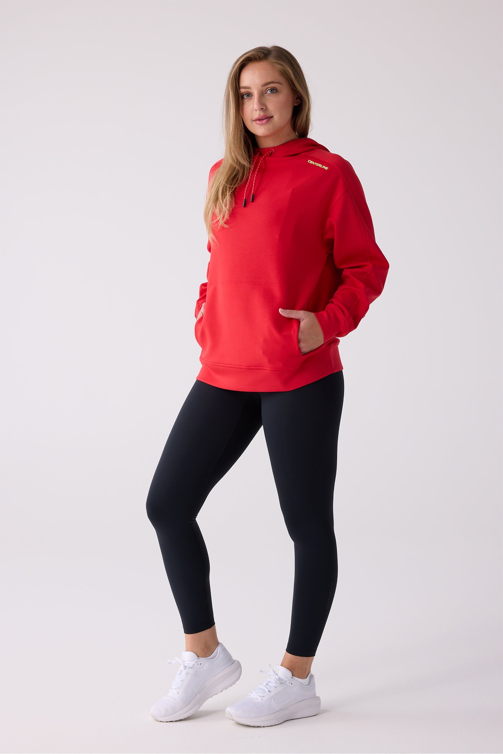 Red hooded sweatshirt by Centerline Athletics with a drawstring hood, front kangaroo pocket, and 'CENTERLINE' text on the shoulder, worn with black leggings and white sneakers.