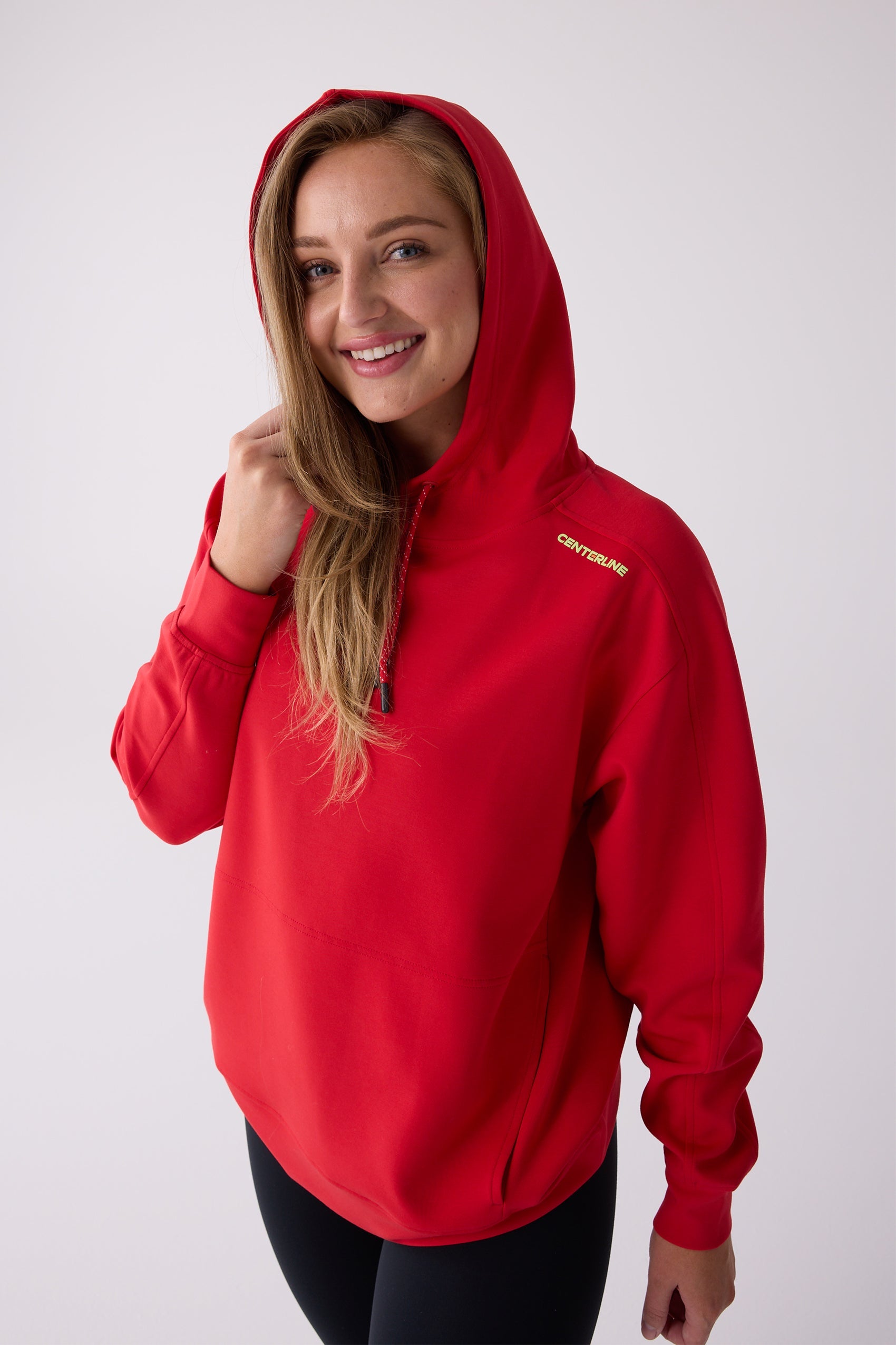 Red Après Women's Hooded Sweatshirt by Centerline Athletics, featuring a yellow CENTERLINE logo, drawstring hood, and front pocket, worn by a smiling woman over black leggings.