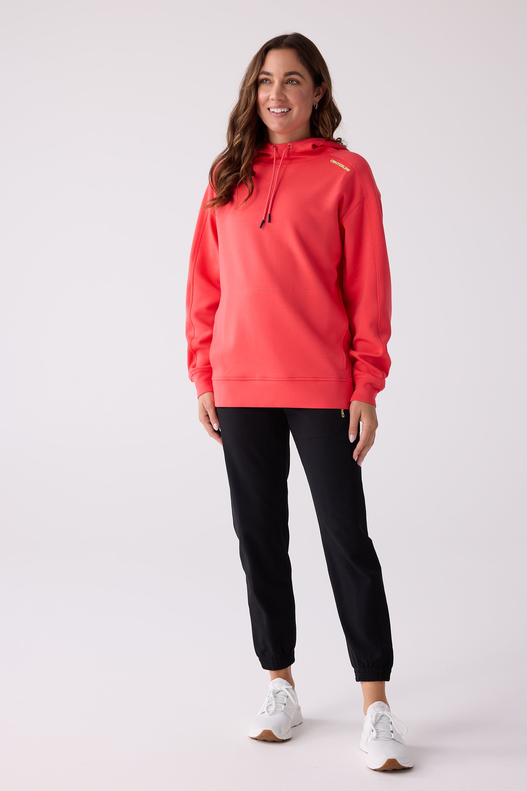 Coral hooded sweatshirt by Centerline Athletics, featuring yellow brand lettering on the shoulder and black drawstring tips, worn with black pants and white sneakers.