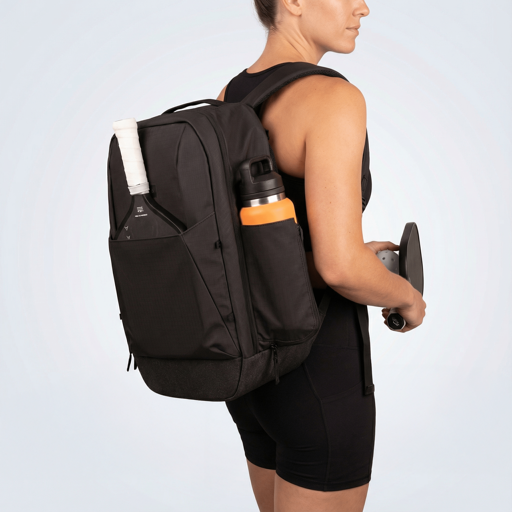 Black pickleball backpack by FORWRD, worn by a woman with a paddle and water bottle in side pockets.