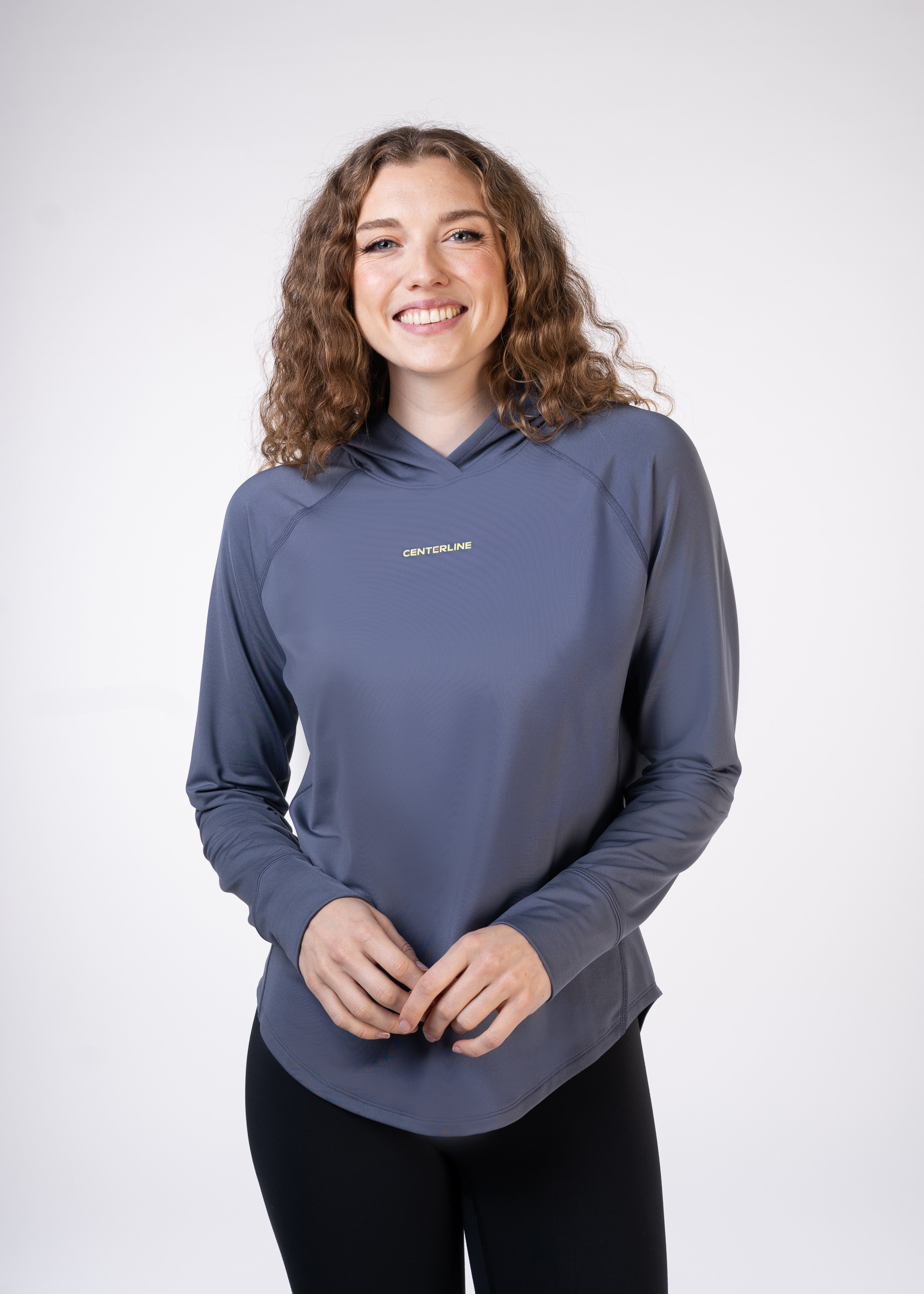 A woman smiles while wearing a long-sleeve gray hooded top and black leggings.