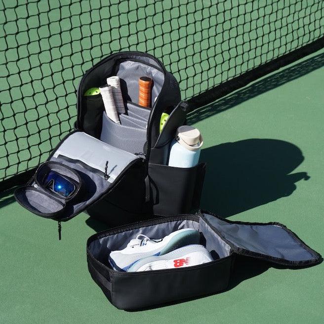 Black pickleball backpack by FORWRD, open on a court with racquets, water bottle, sunglasses, and a separate shoe compartment holding white sneakers.
