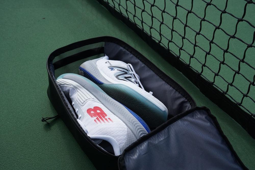 Black shoe cube by FORWRD, unzipped, revealing two white tennis shoes with red and grey logos and colorful soles, on a tennis court with a net.