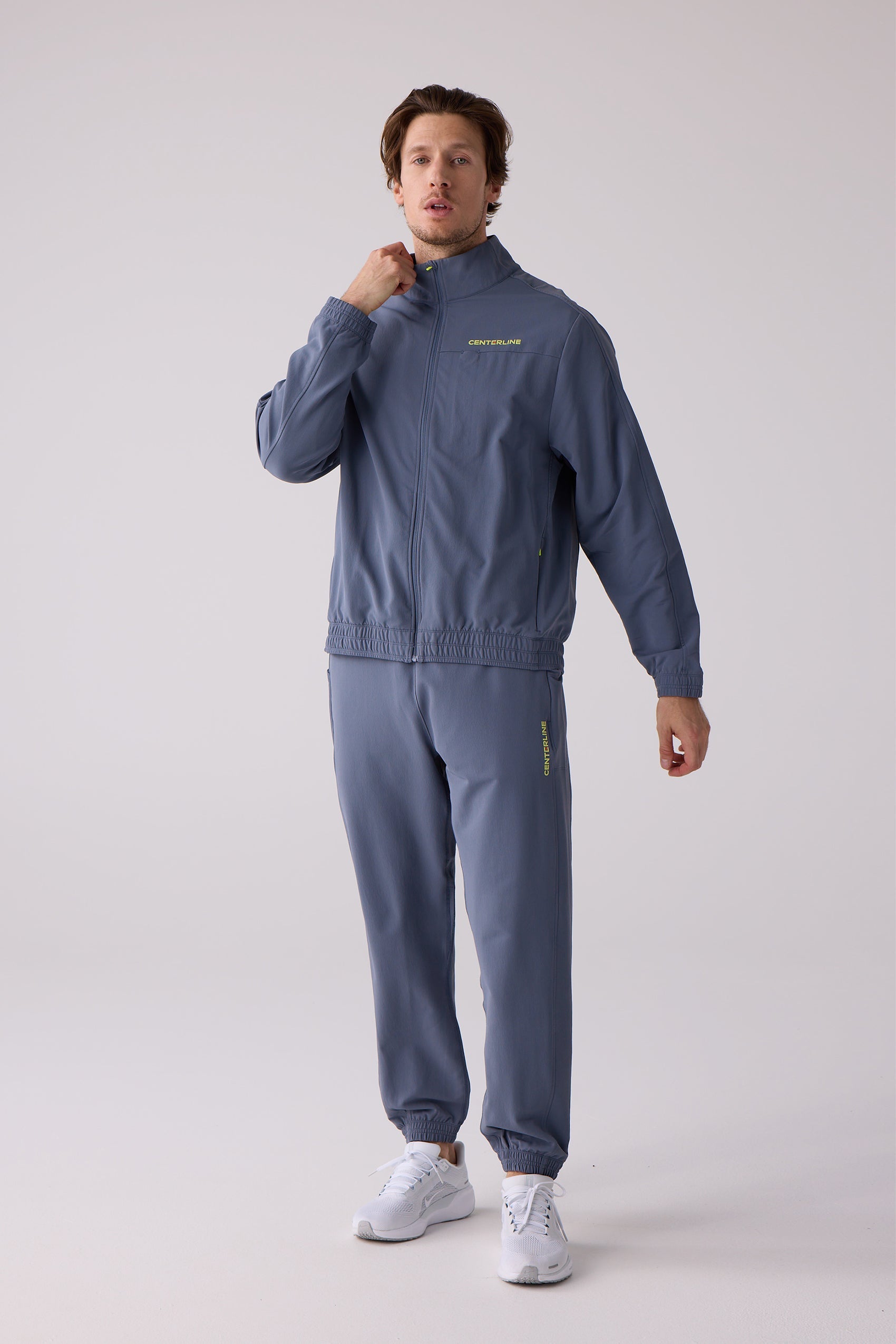 Men's slate blue track jacket by Centerline Athletics with yellow chest logo, worn with matching track pants and white sneakers.