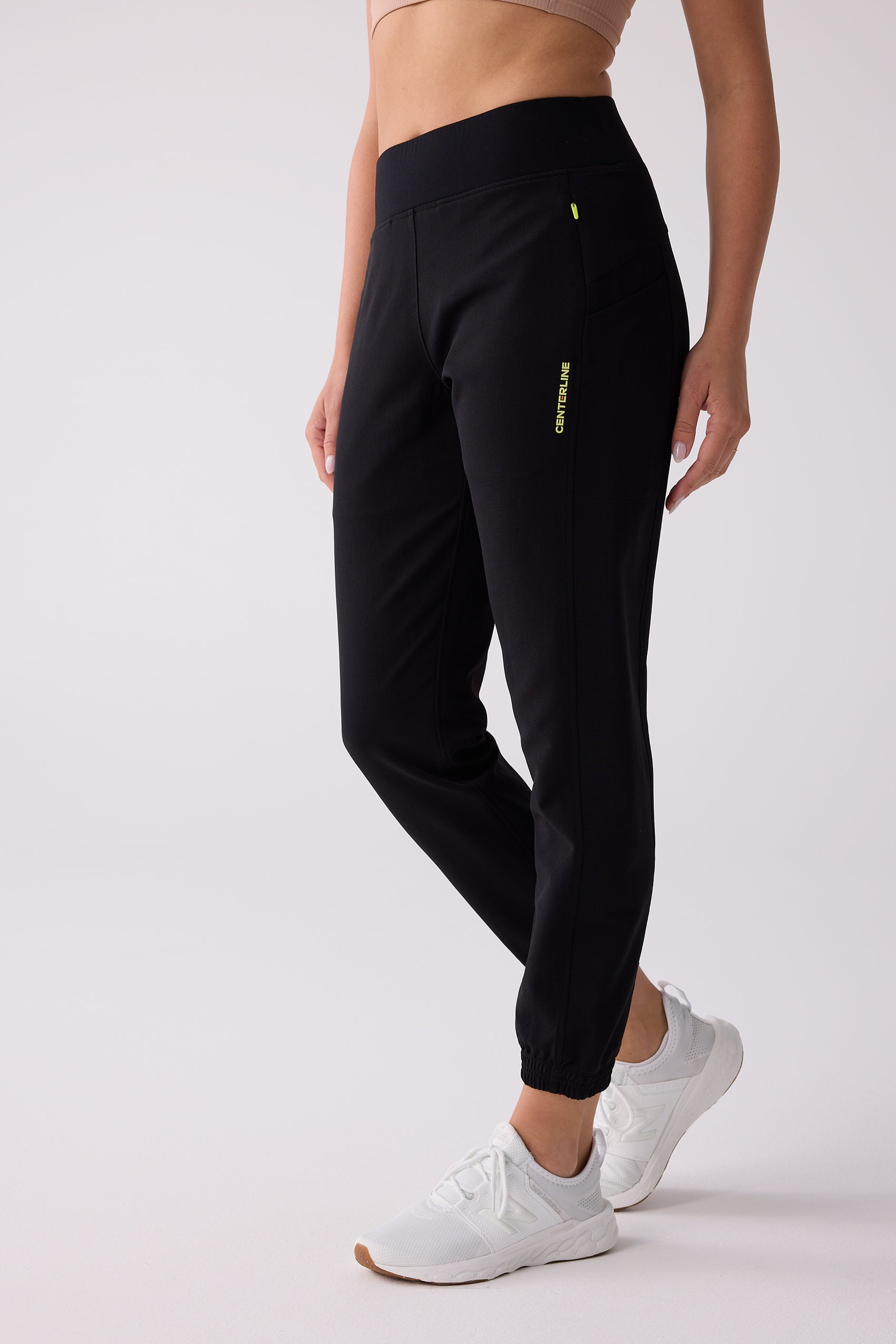Black Women's Courtside Track Pants by Centerline Athletics, with a lime green vertical logo, elasticized cuffs, and paired with white sneakers.