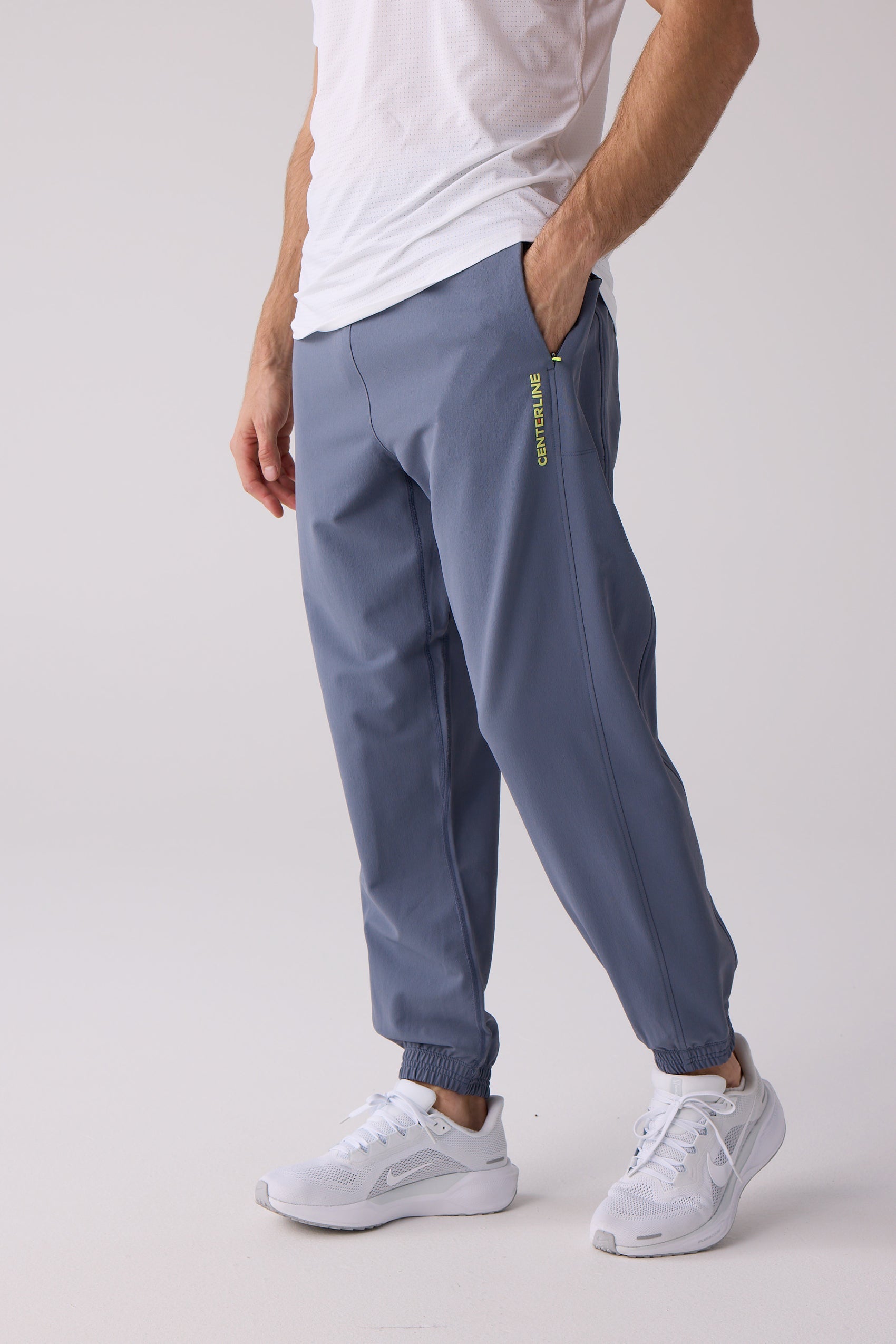 Slate blue Men's Courtside Track Pants by Centerline Athletics, featuring yellow vertical "CENTERLINE" text on the left leg, a yellow side pocket zipper, and cuffed ankles.