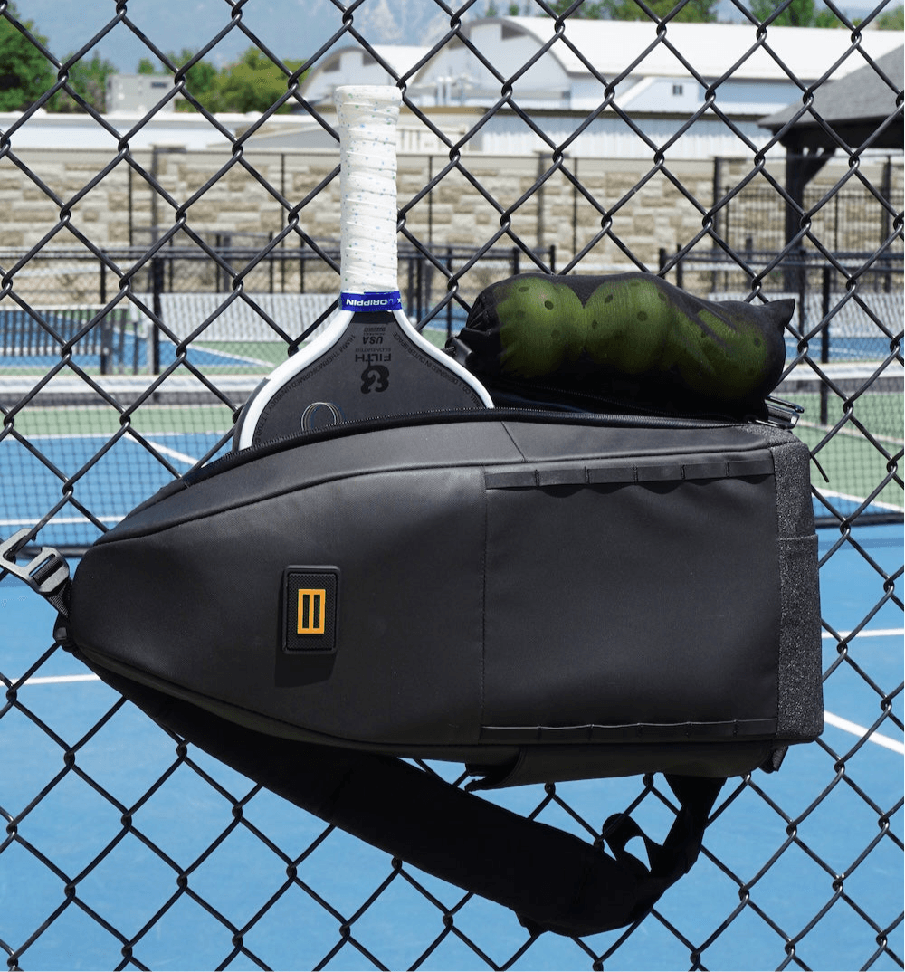 Black pickleball sling bag by FORWRD, with a yellow side logo, containing a white paddle and green pickleballs, hanging on a chain-link fence.