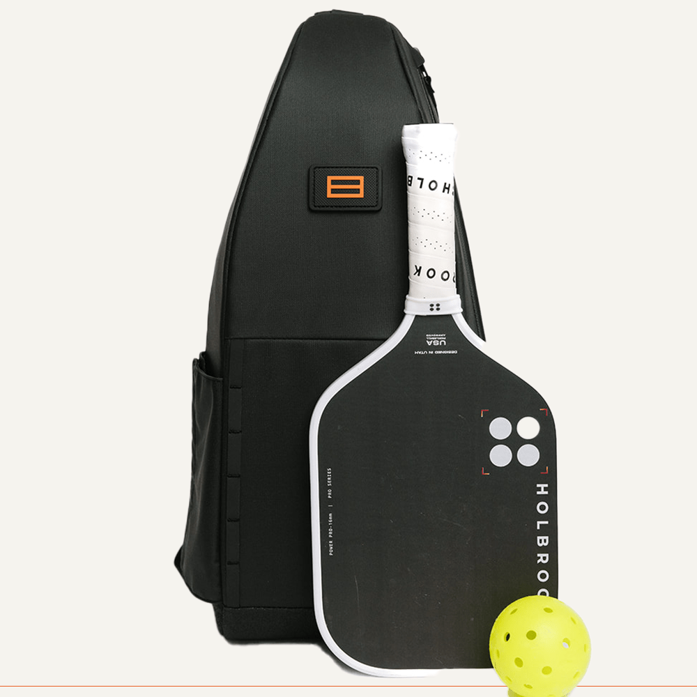 Black pickleball sling bag by FORWRD, standing upright with an orange logo, next to a black and white paddle and a yellow pickleball.