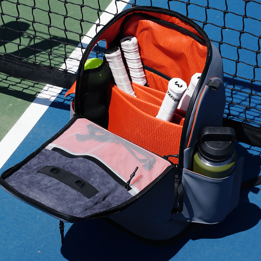 Grey pickleball bag by FORWRD, open on a court displaying white racket grips, balls, and a water bottle.