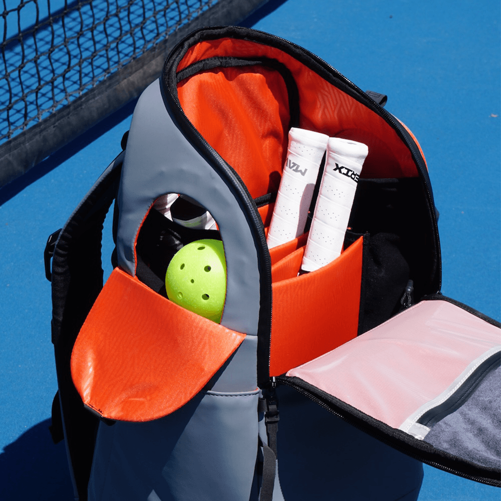 Grey and orange Court Caddy Pickleball Bag by FORWRD, open with a lime green pickleball, white paddles, and a mesh pocket on a blue court.