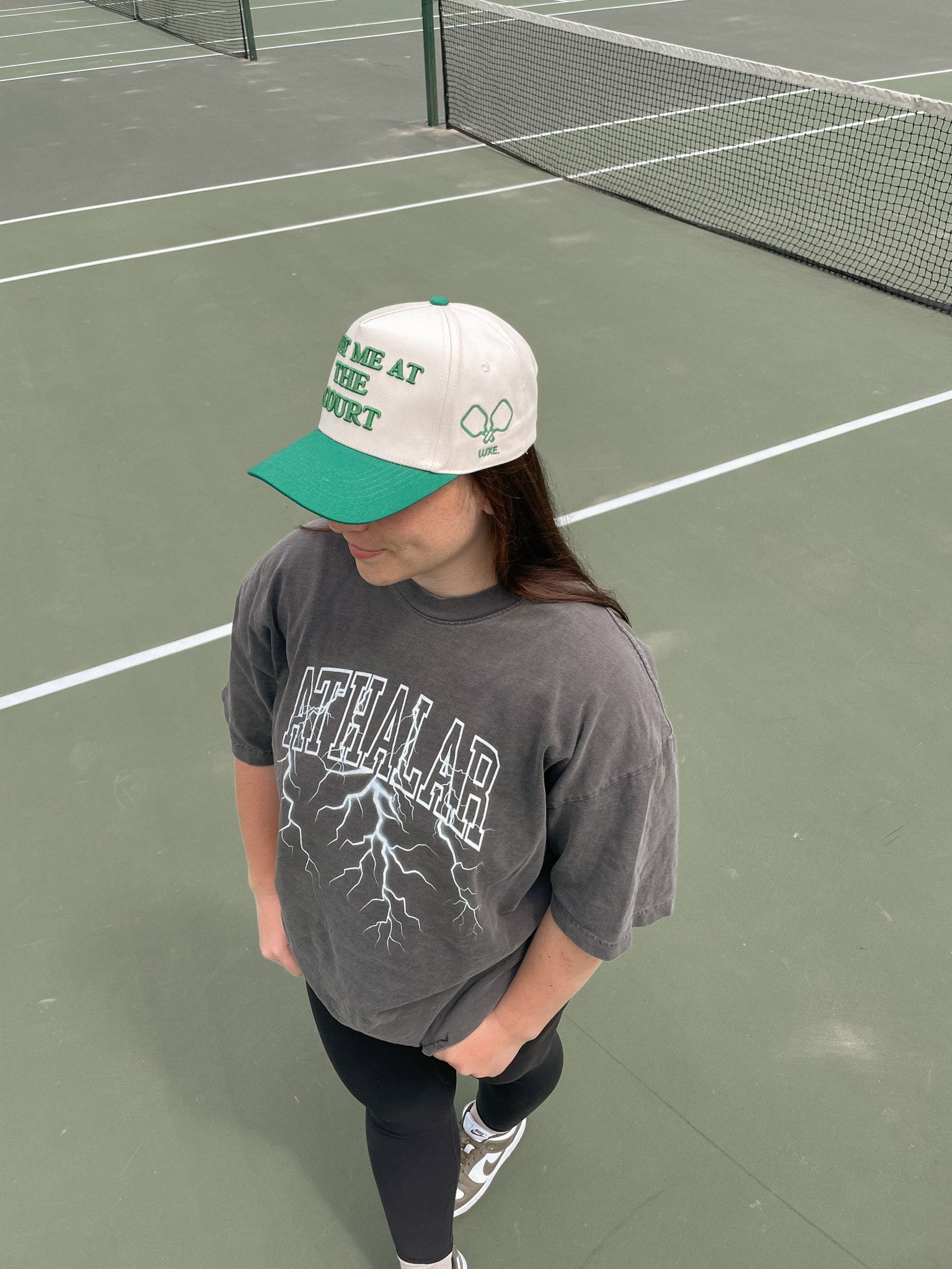 Overhead view of a person wearing a beige and green baseball cap with the phrase "ME AT THE COURT" and a gray graphic t-shirt with lightning bolts on a tennis court.