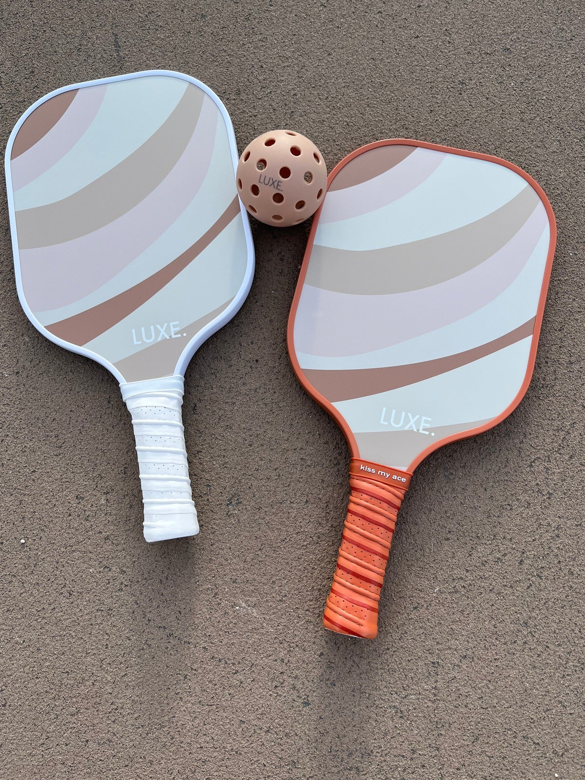 Two pickleball paddles and a ball on pavement. One paddle is white, one is orange, both feature a neutral-toned wave design.