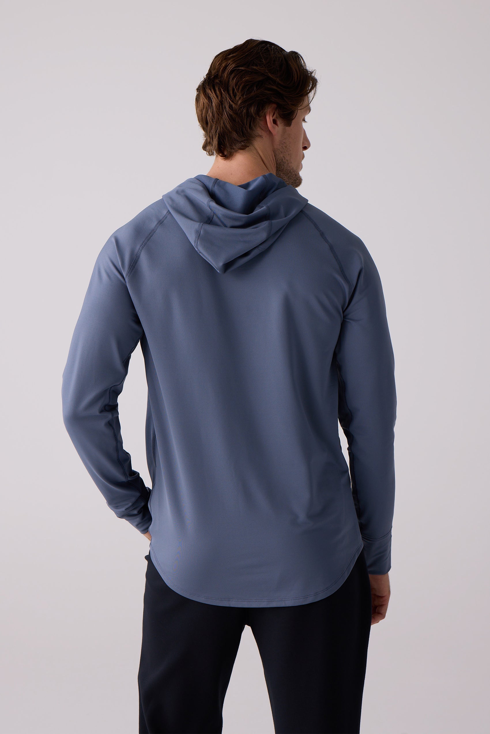 Blue men's layering hoodie by Centerline Athletics, back view, with a hood, raglan sleeves, and curved hem, worn with black pants.