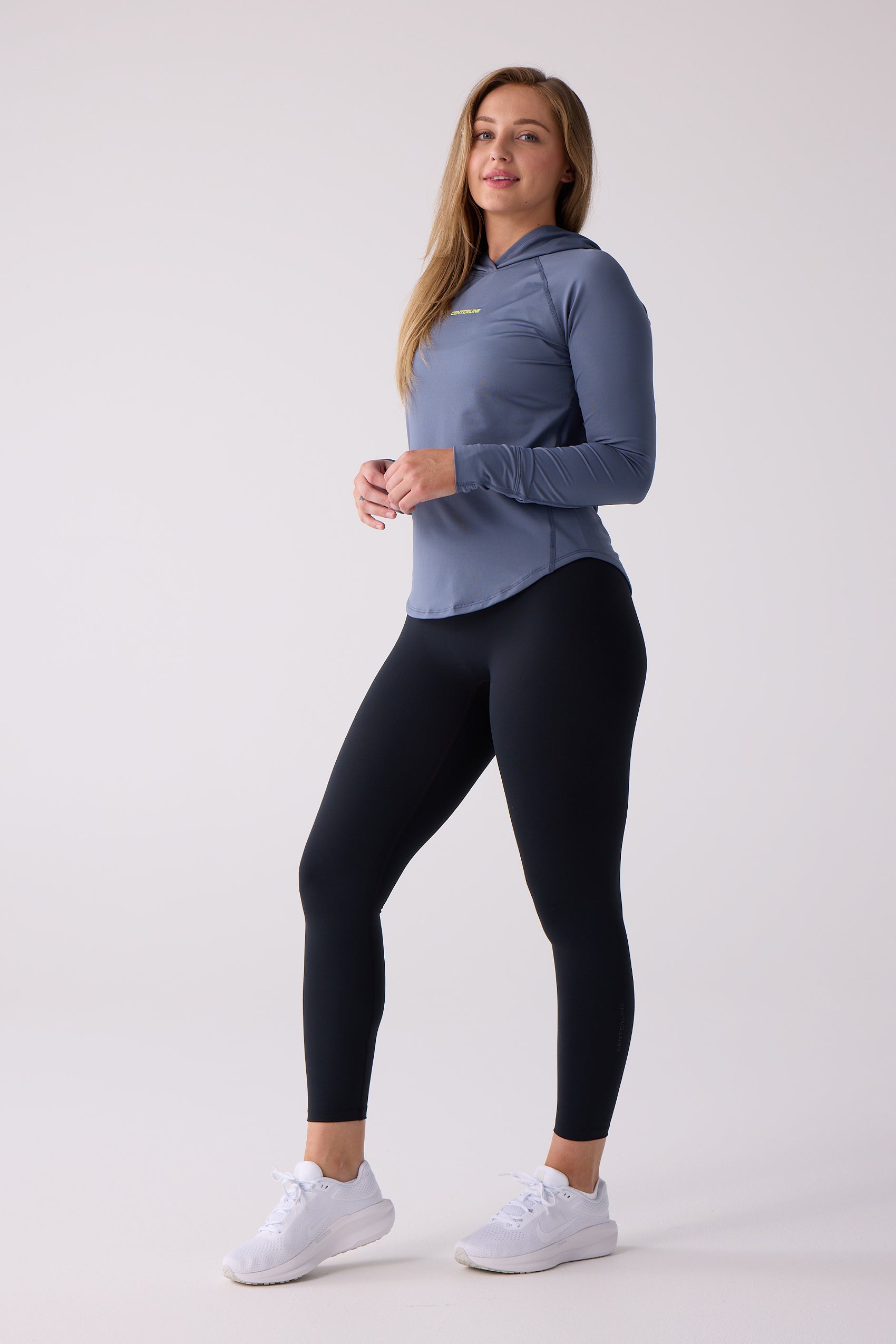 Blue-gray long-sleeved women's layering hoodie by Centerline Athletics, with a hood, rounded hem, and raglan sleeves, worn with black leggings and white sneakers.