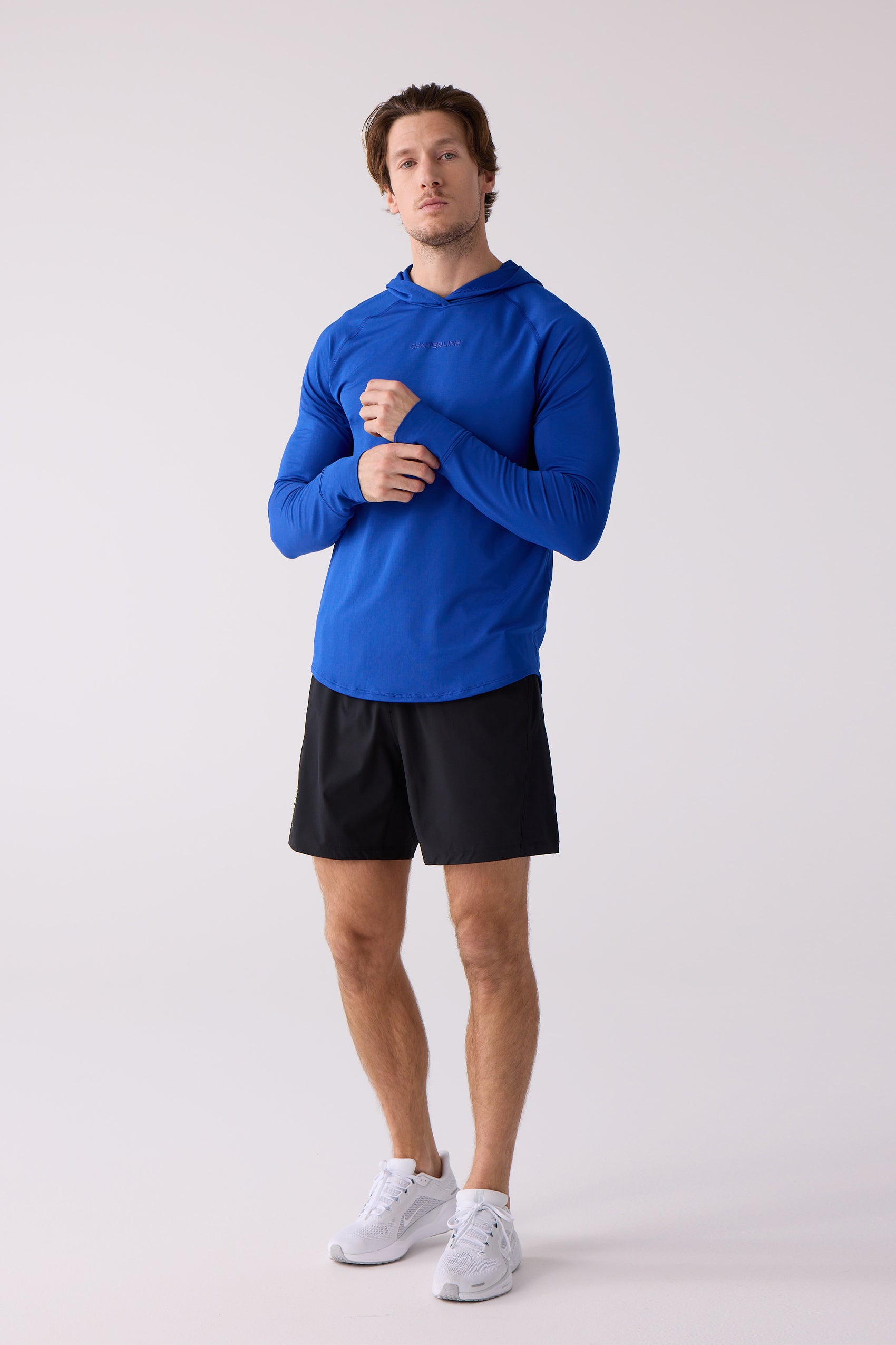 Men's blue layering hoodie by Centerline Athletics with logo on chest, worn by a man with black shorts and white sneakers, adjusting the sleeve.