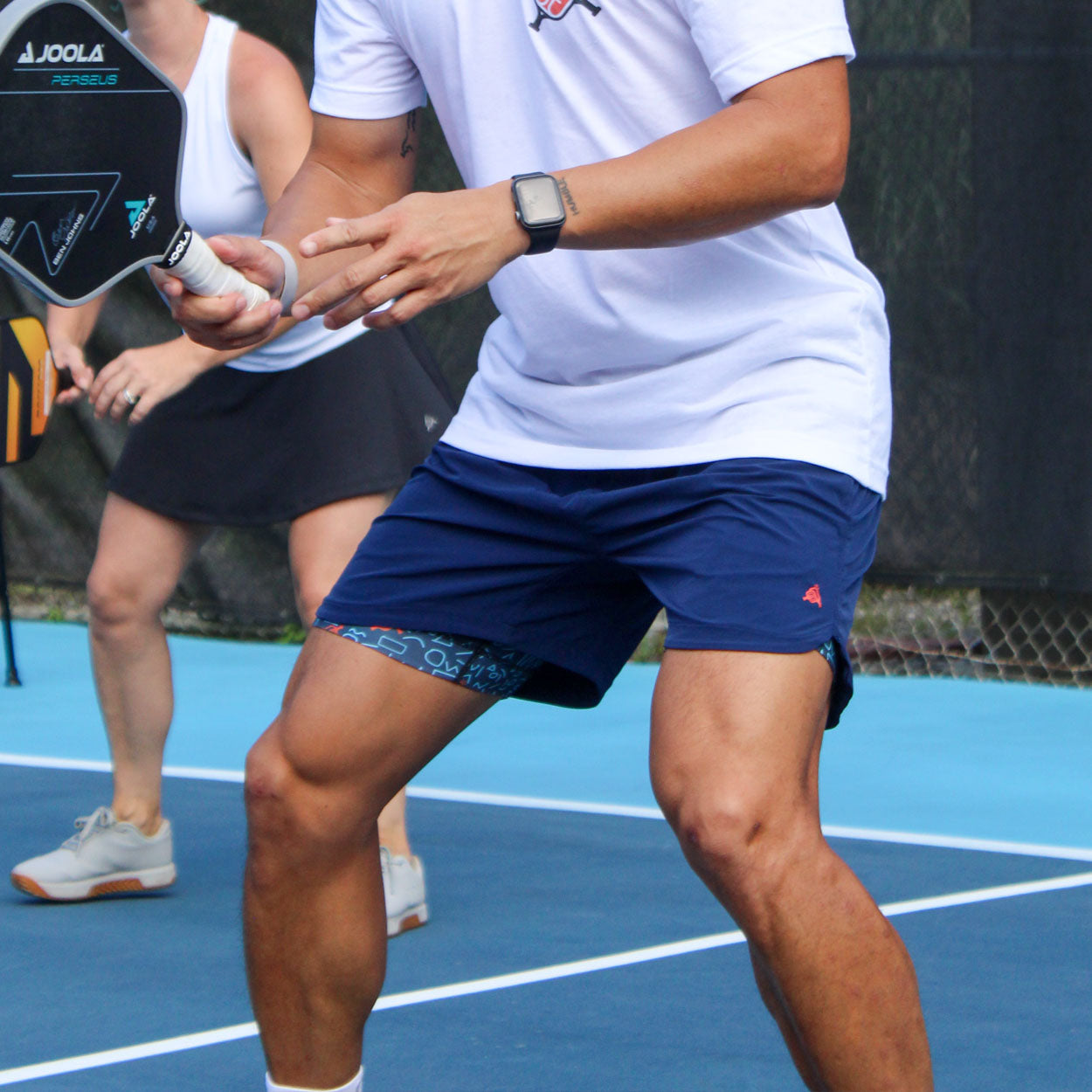 Blue men's pickleball shorts by Good Get with a red rooster logo, worn over geometric patterned compression shorts on a blue court.