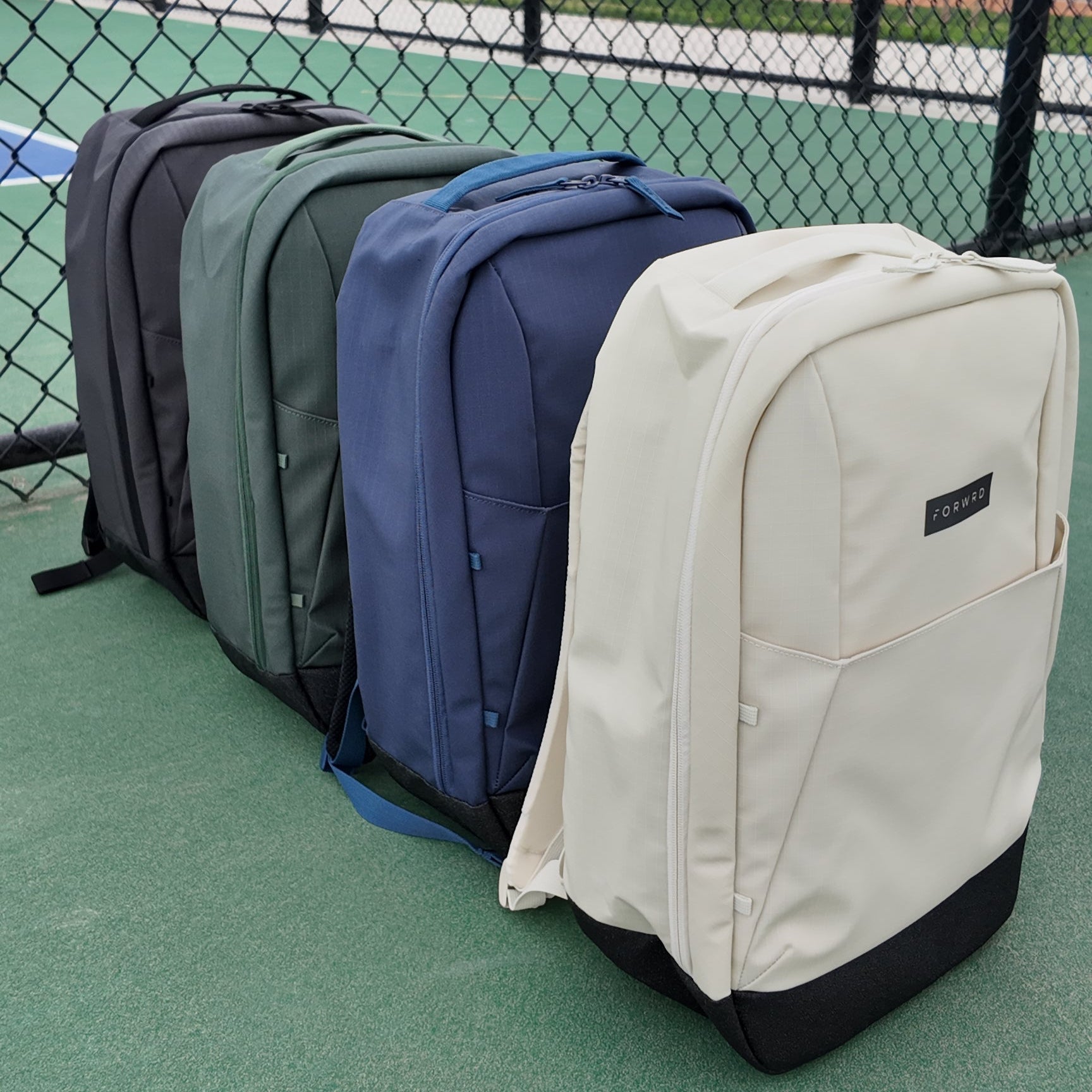 Four Pickleball Backpacks by FORWRD in cream, navy, green, and grey, lined on a court with top handles and side pockets.