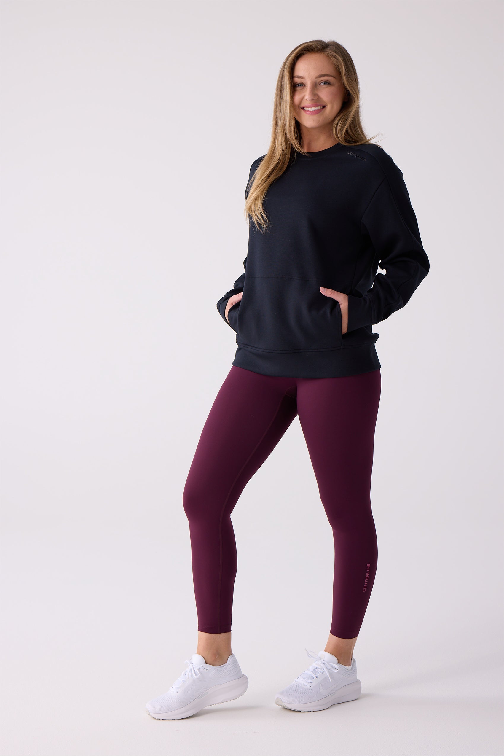 Deep plum 7/8 leggings by Centerline Athletics, worn by a smiling woman with a navy sweatshirt and white sneakers, featuring a subtle leg logo.