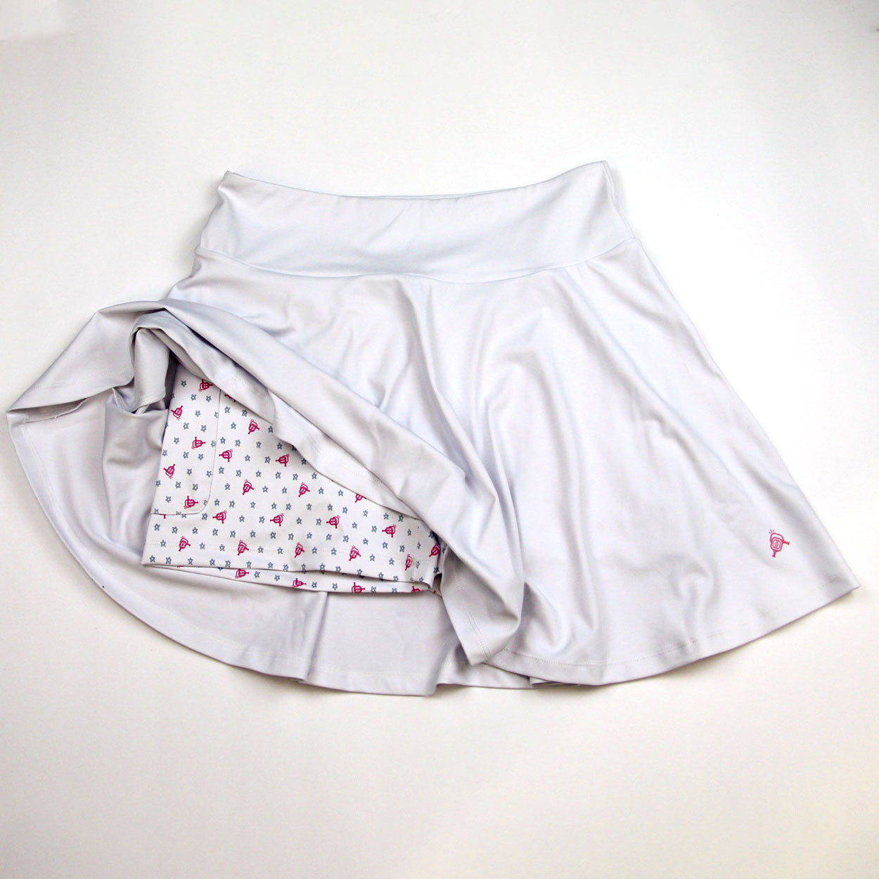 Light grey athletic skirt by Good Get, revealing built-in shorts with a pink bell and star pattern and a small pink logo.